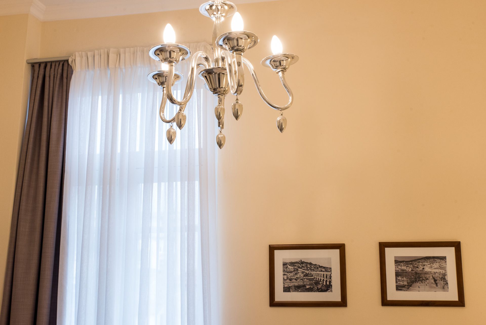 Ένας κρυστάλλινος πολυέλαιος κρέμεται πάνω από ένα παράθυρο με πολυεπίπεδες λευκές και γκρι κουρτίνες, δίπλα σε δύο πλαισιωμένες εκτυπώσεις σε έναν κρεμ τοίχο.