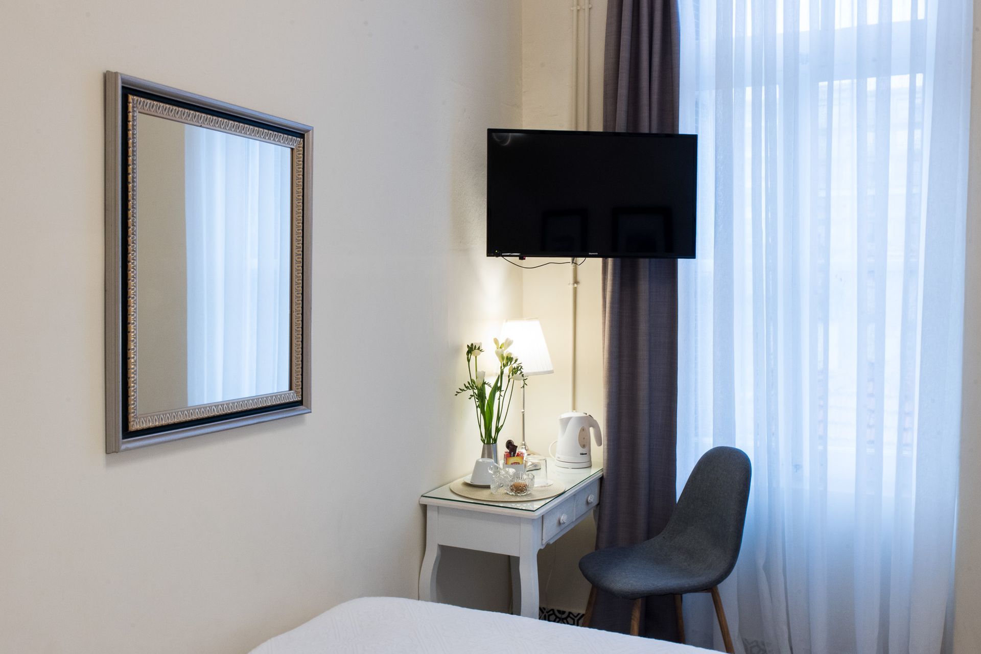 Μια γωνιά δωματίου ξενοδοχείου με έναν καθρέφτη τοίχου, ένα λευκό γραφείο, μια μικρή λάμπα, μια τηλεόραση και μια γκρι καρέκλα δίπλα σε ένα παράθυρο με κουρτίνες.