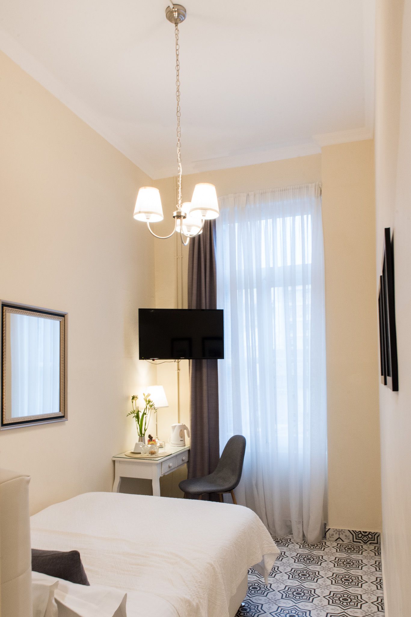 Ένα φωτεινό δωμάτιο ξενοδοχείου με ένα κρεβάτι, ένα μικρό γραφείο, μια εντοιχισμένη τηλεόραση, έναν πολυέλαιο και ένα δάπεδο με πλακάκια με σχέδια.