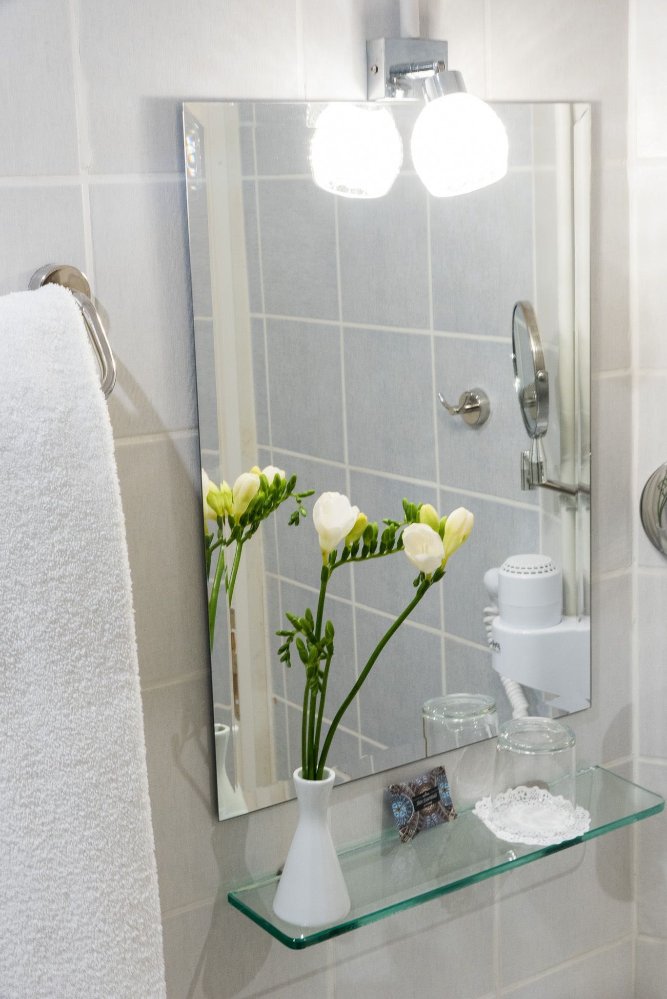 Ένα νιπτήρα μπάνιου με καθρέφτη, ένα γυάλινο ράφι που περιέχει ένα λευκό βάζο με λουλούδια και μια λευκή πετσέτα που κρέμεται στα αριστερά.