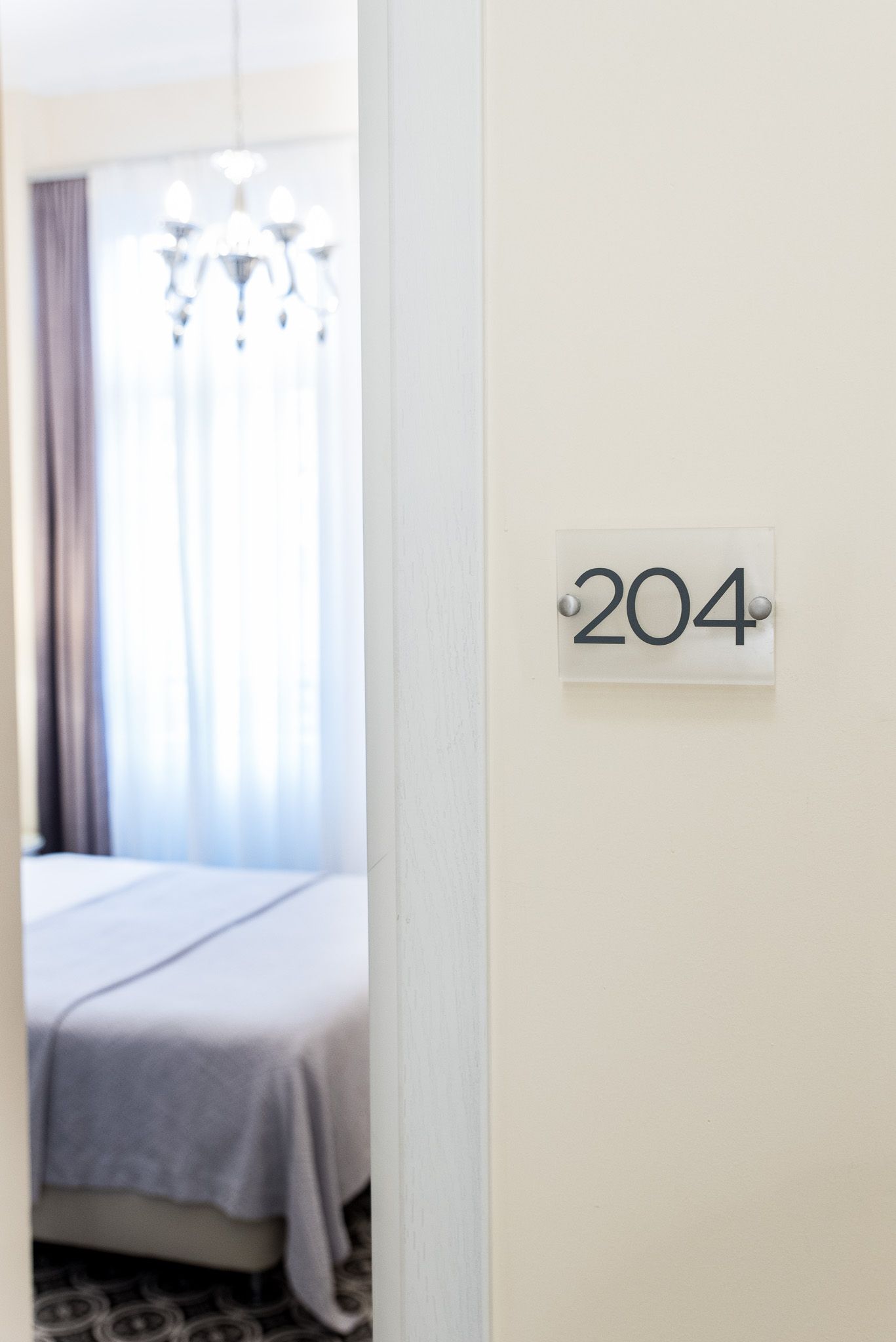 Μια πόρτα δωματίου ξενοδοχείου με μια ευκρινή πινακίδα που υποδεικνύει τον αριθμό δωματίου 204, η οποία δείχνει ένα υπνοδωμάτιο με κρεβάτι και πολυέλαιο στο εσωτερικό.