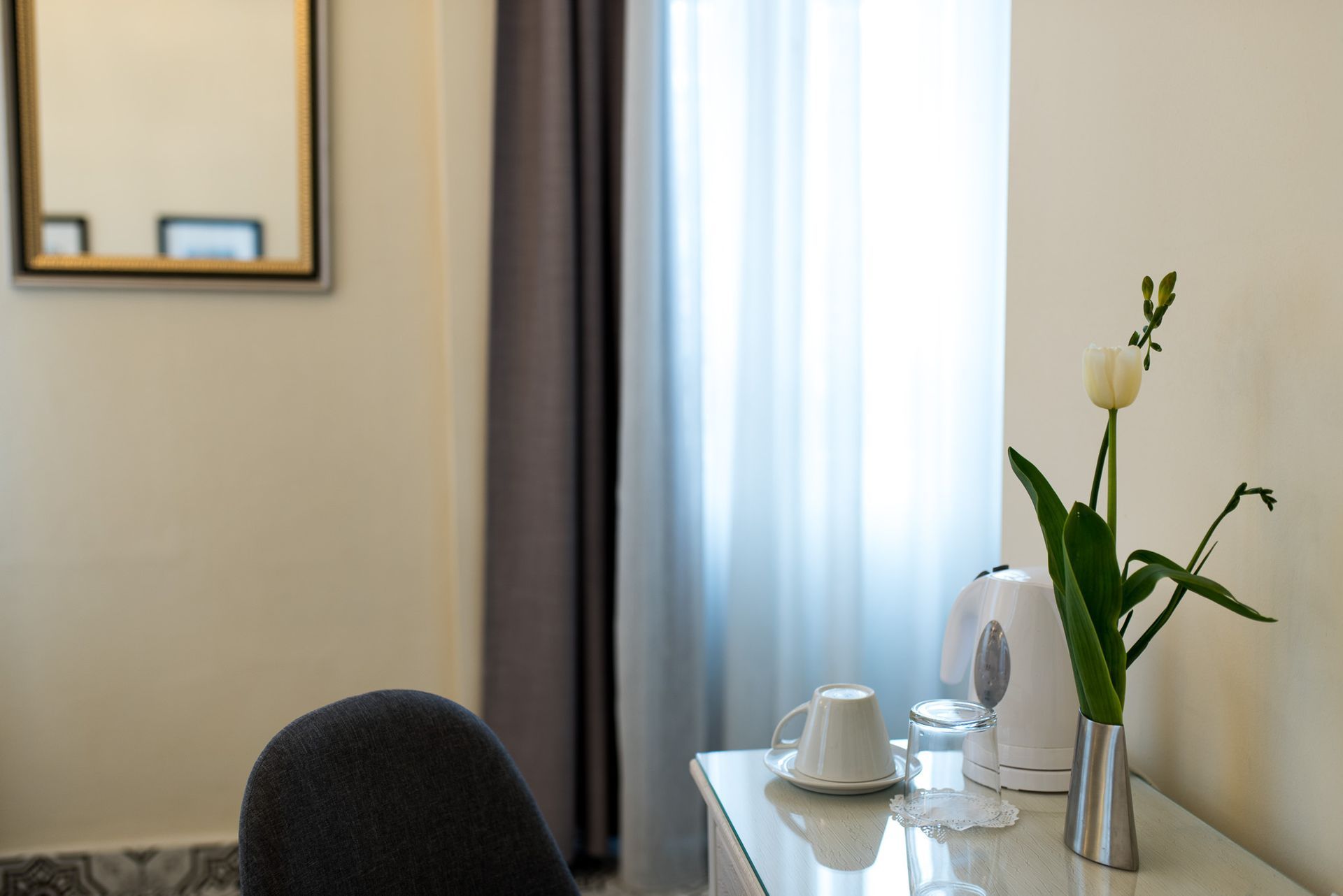 Μια λευκή βραστήρας, ένα φλιτζάνι σε ένα πιατάκι και ένα λευκό λουλούδι σε ένα ασημένιο βάζο βρίσκονται σε ένα λευκό τραπέζι κοντά σε ένα παράθυρο με κουρτίνες.
