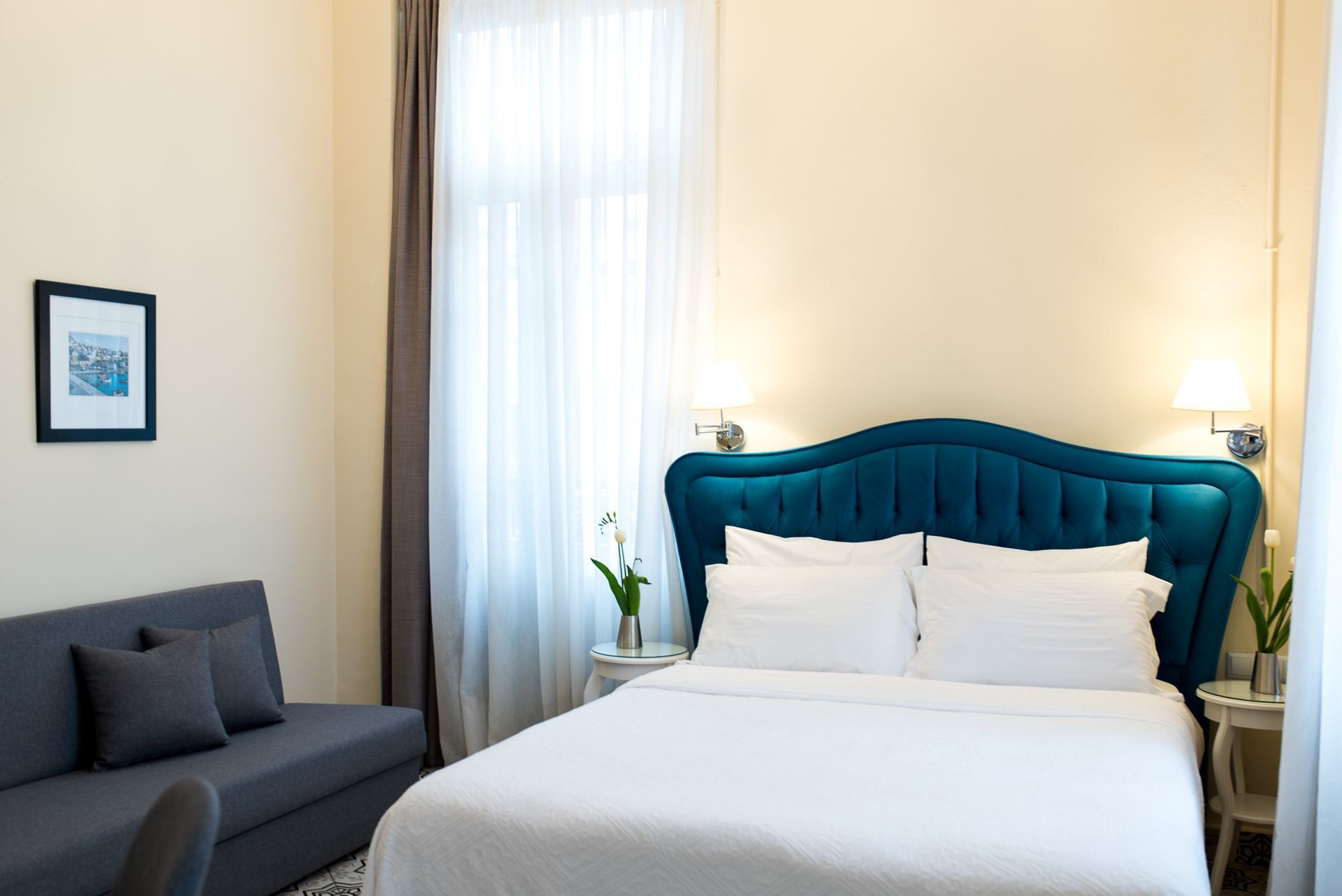 Ένα δωμάτιο ξενοδοχείου με λευκό κρεβάτι, γαλαζοπράσινο κεφαλάρι με φουντωτά καλύμματα, γκρι καναπέ, παράθυρο με διαφανείς κουρτίνες και πλαισιωμένη τέχνη τοίχου.