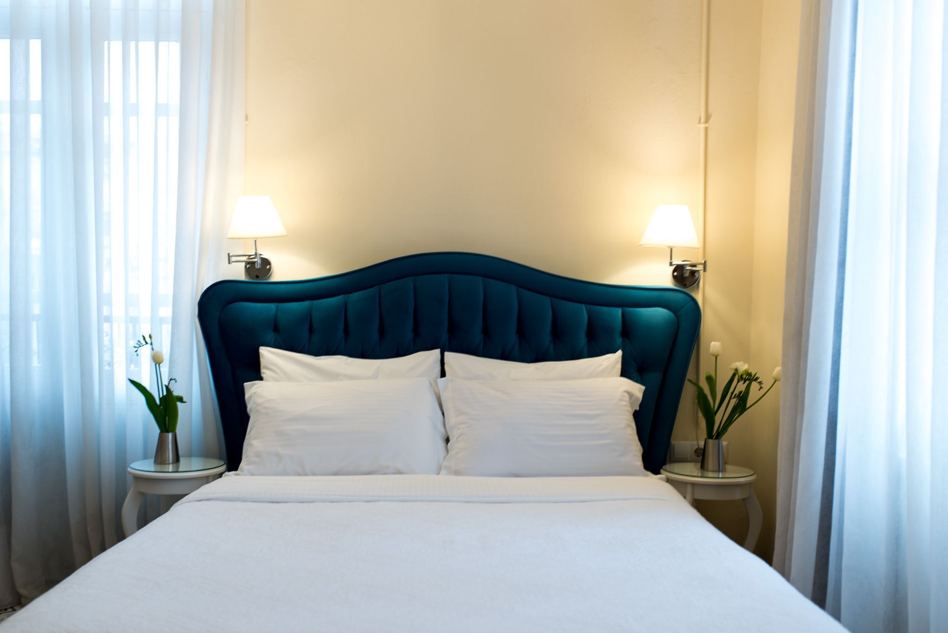 Ένα στρωμένο κρεβάτι με γαλαζοπράσινο κεφαλάρι με φουντωτά υφάσματα, λευκά σεντόνια και κομοδίνα με λουλούδια, πλαισιωμένα από λευκές κουρτίνες.