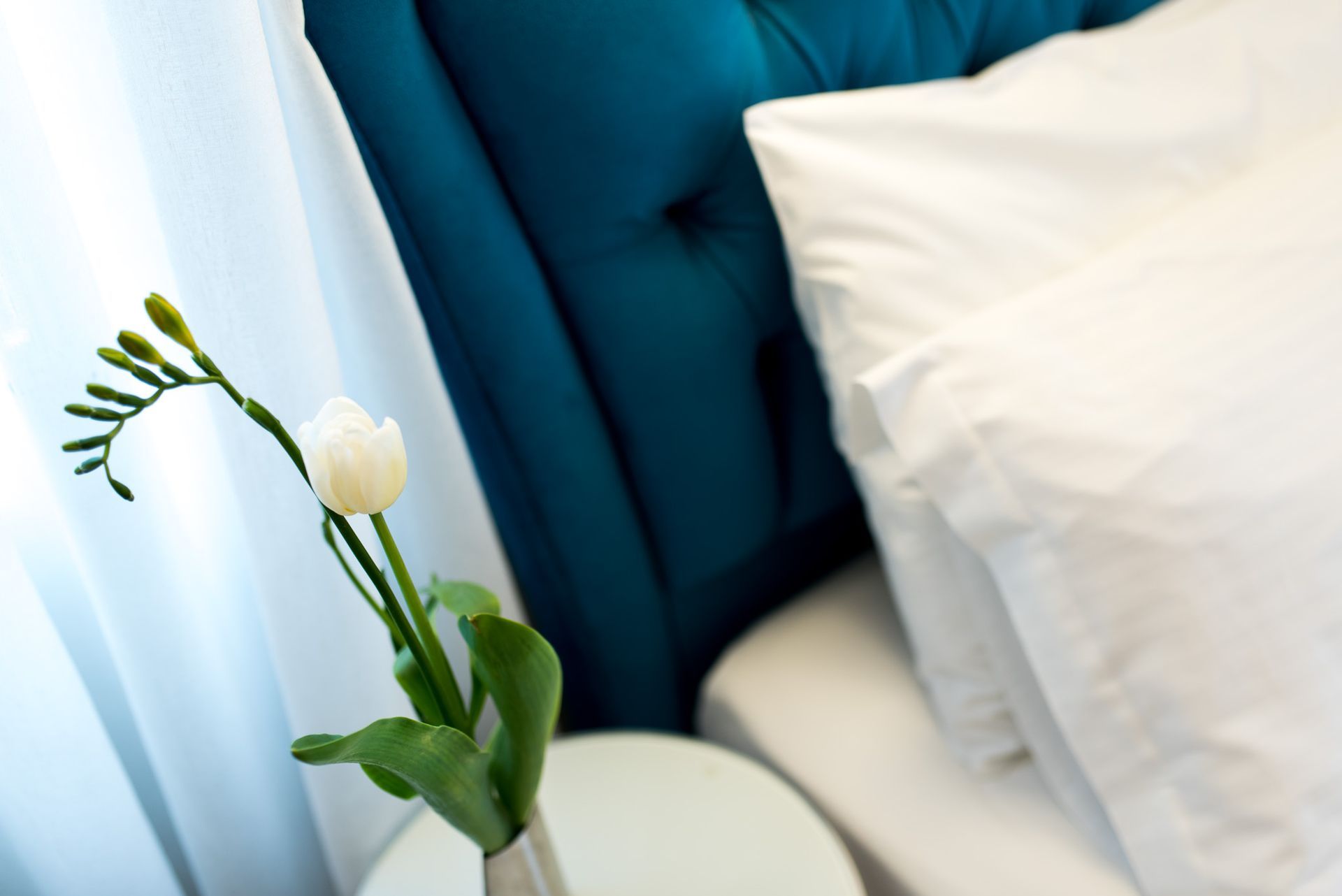 Ένα λευκό λουλούδι σε ένα βάζο κάθεται σε ένα στρογγυλό τραπέζι δίπλα σε ένα κρεβάτι με κεφαλάρι σε χρώμα γαλαζοπράσινου φουντωτού και λευκά μαξιλάρια.