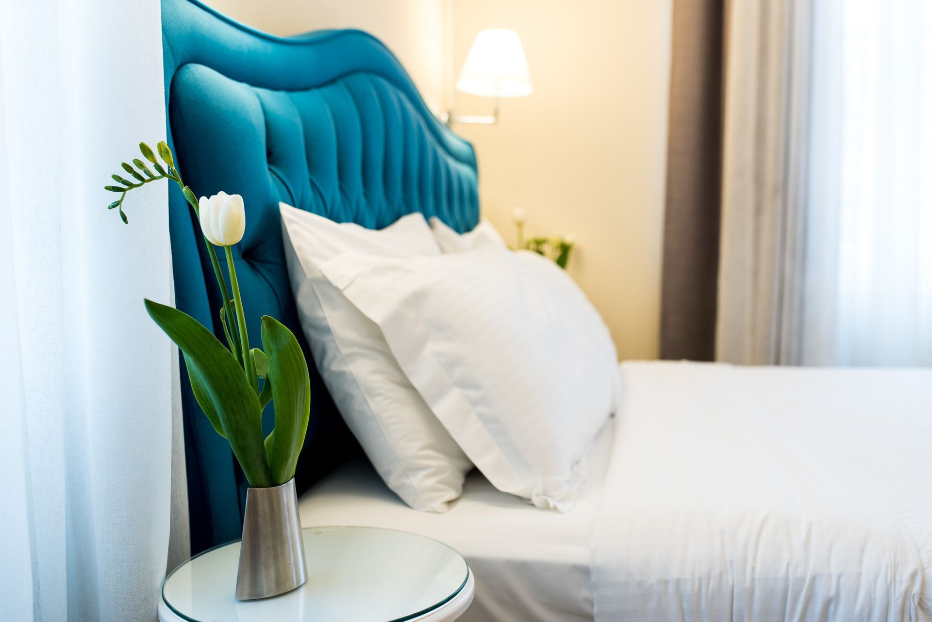 Ένα κομοδίνο με ένα λευκό λουλούδι σε βάζο δίπλα σε ένα κρεβάτι με λευκά μαξιλάρια και ένα γαλαζοπράσινο κεφαλάρι με φουντωτά κεντήματα.