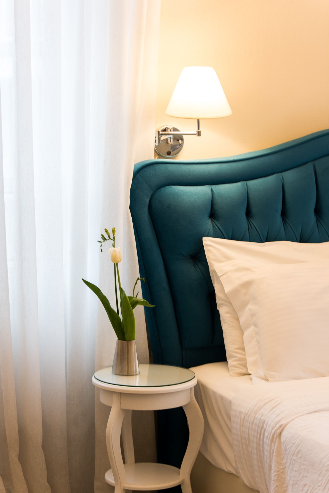 Ένα λευκό κομοδίνο με ένα μικρό φυτό δίπλα σε ένα γαλαζοπράσινο κεφαλάρι με κουμπιά και ένα λευκό απλίκα τοίχου από πάνω.