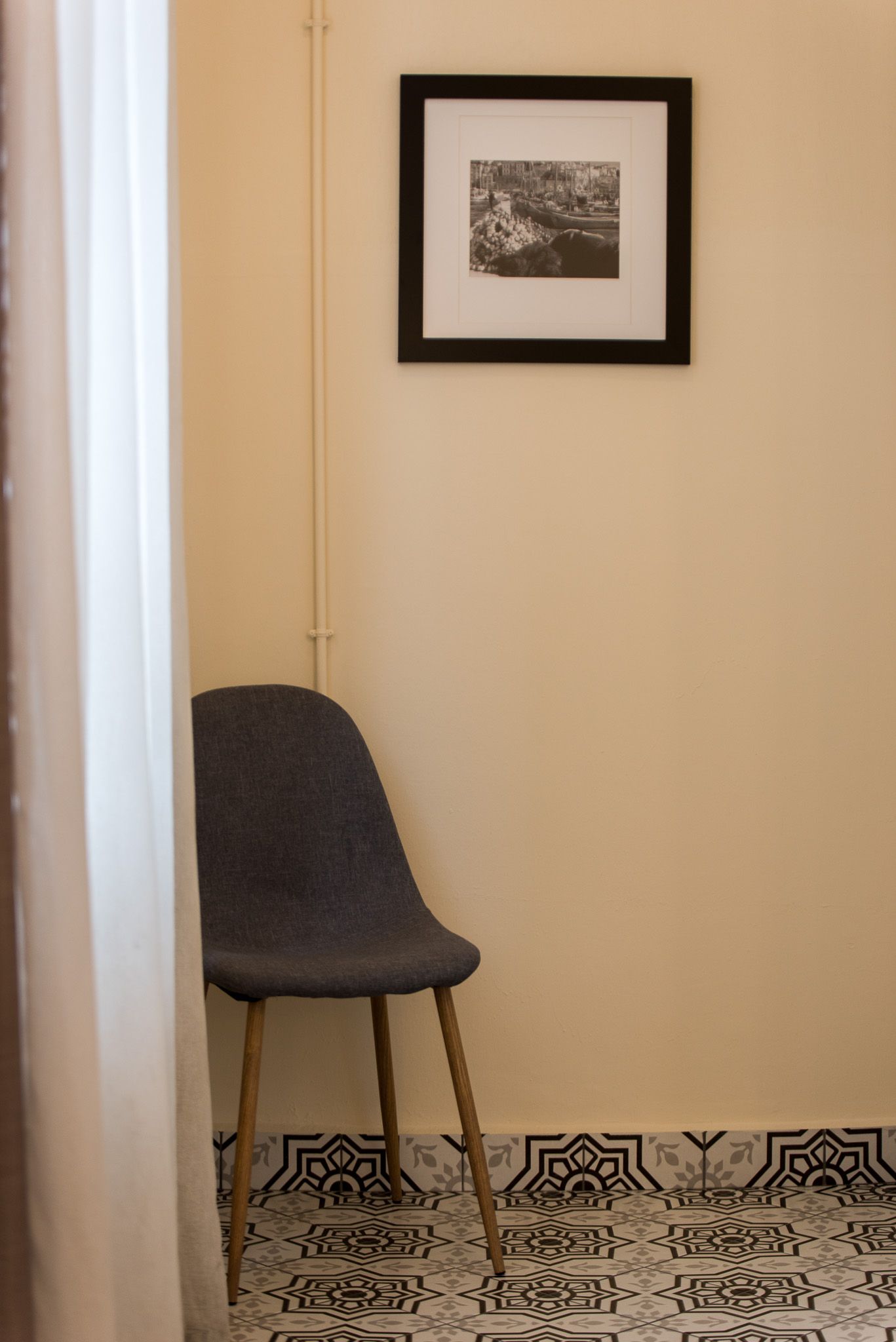 Μια σκούρα γκρι καρέκλα κάθεται σε μια γωνία δίπλα σε έναν μπεζ τοίχο, με μια πλαισιωμένη εικόνα από πάνω και πλακάκια δαπέδου με σχέδια από κάτω.