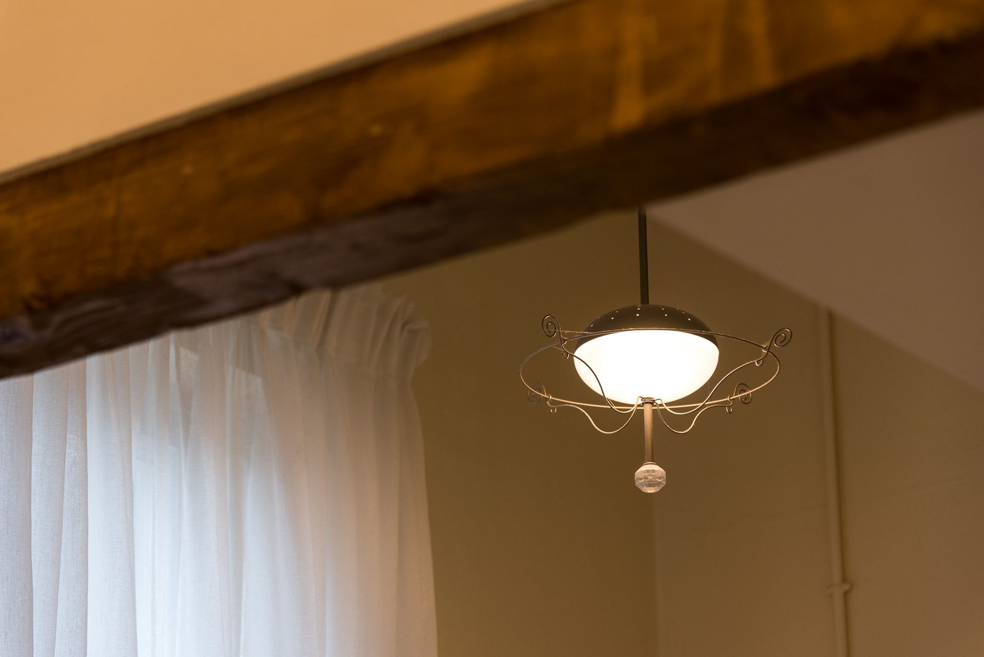 Ένα φωτιζόμενο διακοσμητικό κρεμαστό φωτιστικό με περίπλοκα μεταλλικά κλαδιά κρέμεται κοντά σε λευκές κουρτίνες πίσω από μια ρουστίκ ξύλινη δοκό.