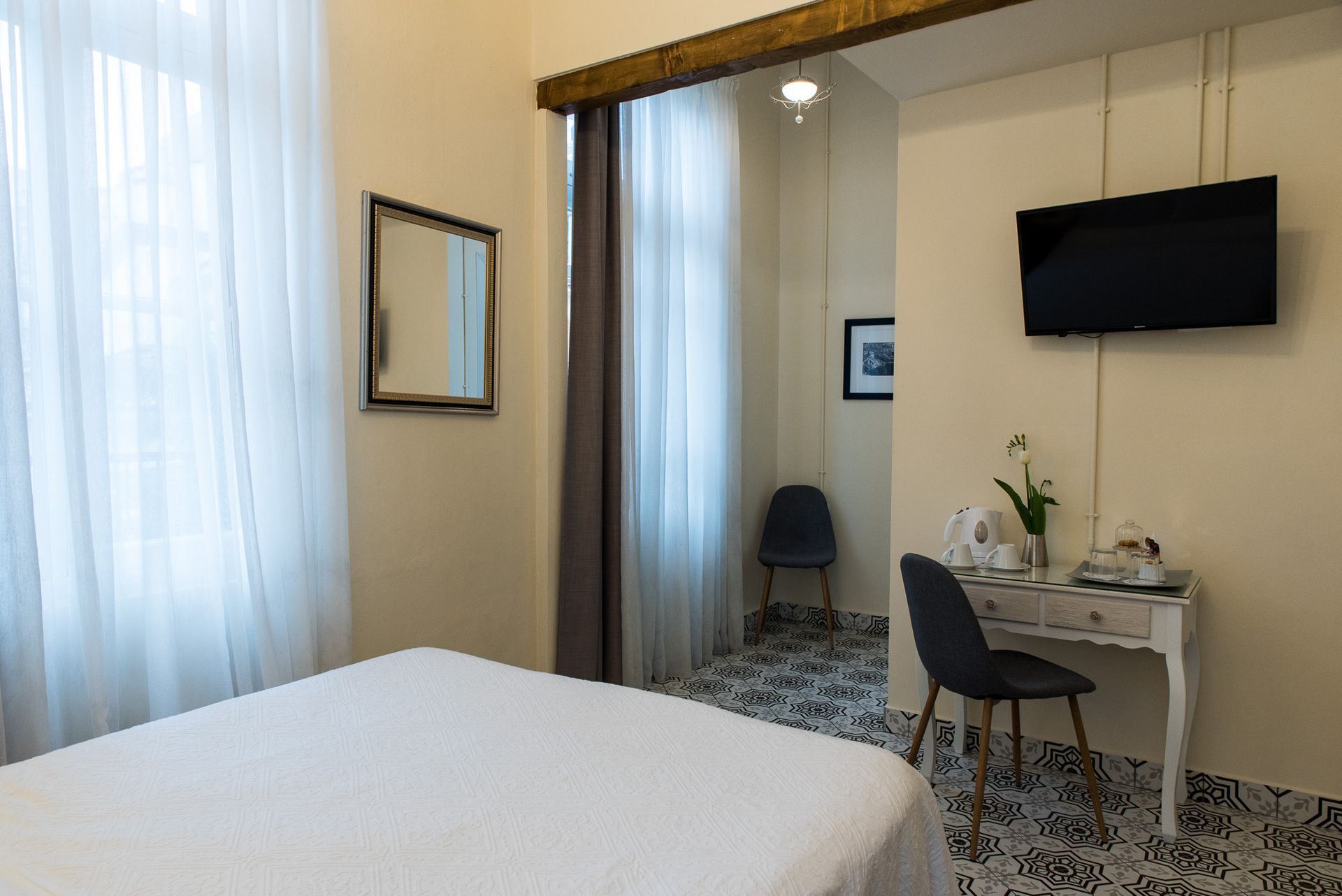 Ένα δωμάτιο ξενοδοχείου με ένα κρεβάτι, ένα γραφείο με τηλεόραση από πάνω και ένα μικρό καθιστικό δίπλα σε ένα παράθυρο με λευκές κουρτίνες.