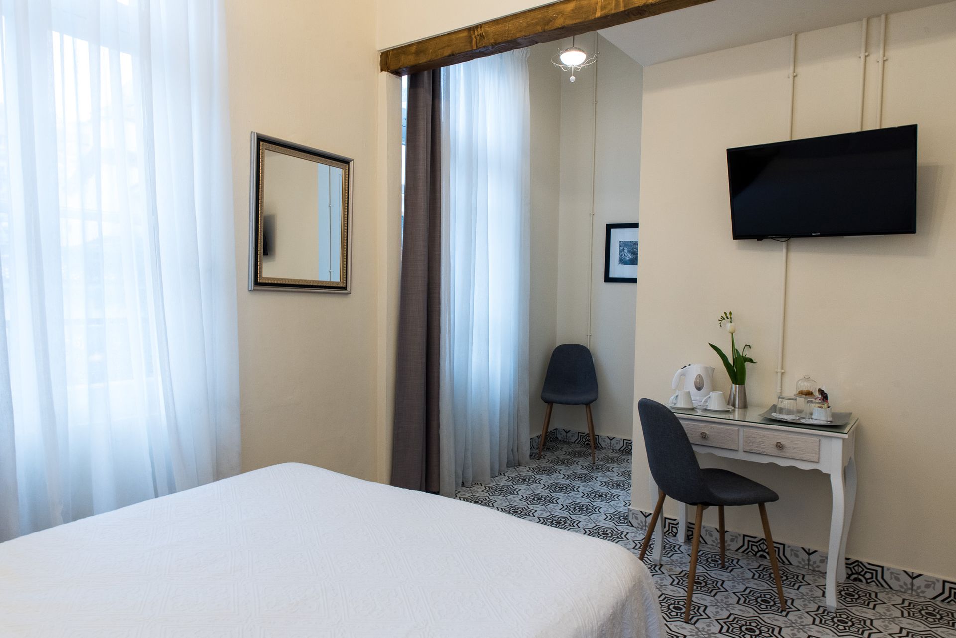 Ένα δωμάτιο ξενοδοχείου με ένα κρεβάτι, ένα γραφείο, μια τηλεόραση, έναν καθρέφτη και μια μικρή καρέκλα δίπλα σε ένα παράθυρο με λευκές κουρτίνες.