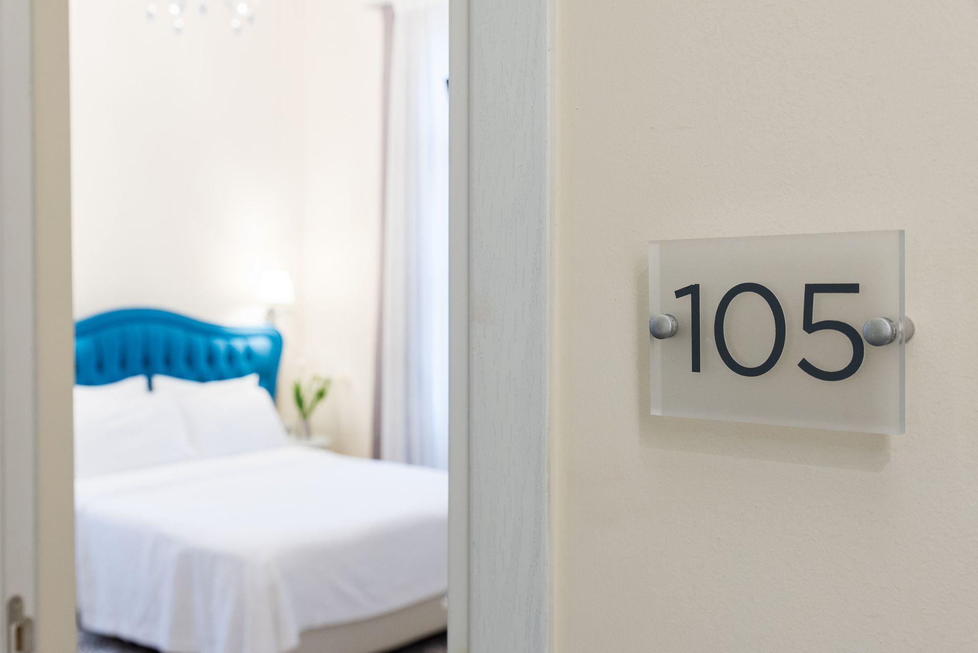 Μια επιτοίχια πινακίδα με την ένδειξη «105» δίπλα σε μια πόρτα, που παρέχει μερική θέα σε ένα δωμάτιο ξενοδοχείου με μπλε κεφαλάρι.