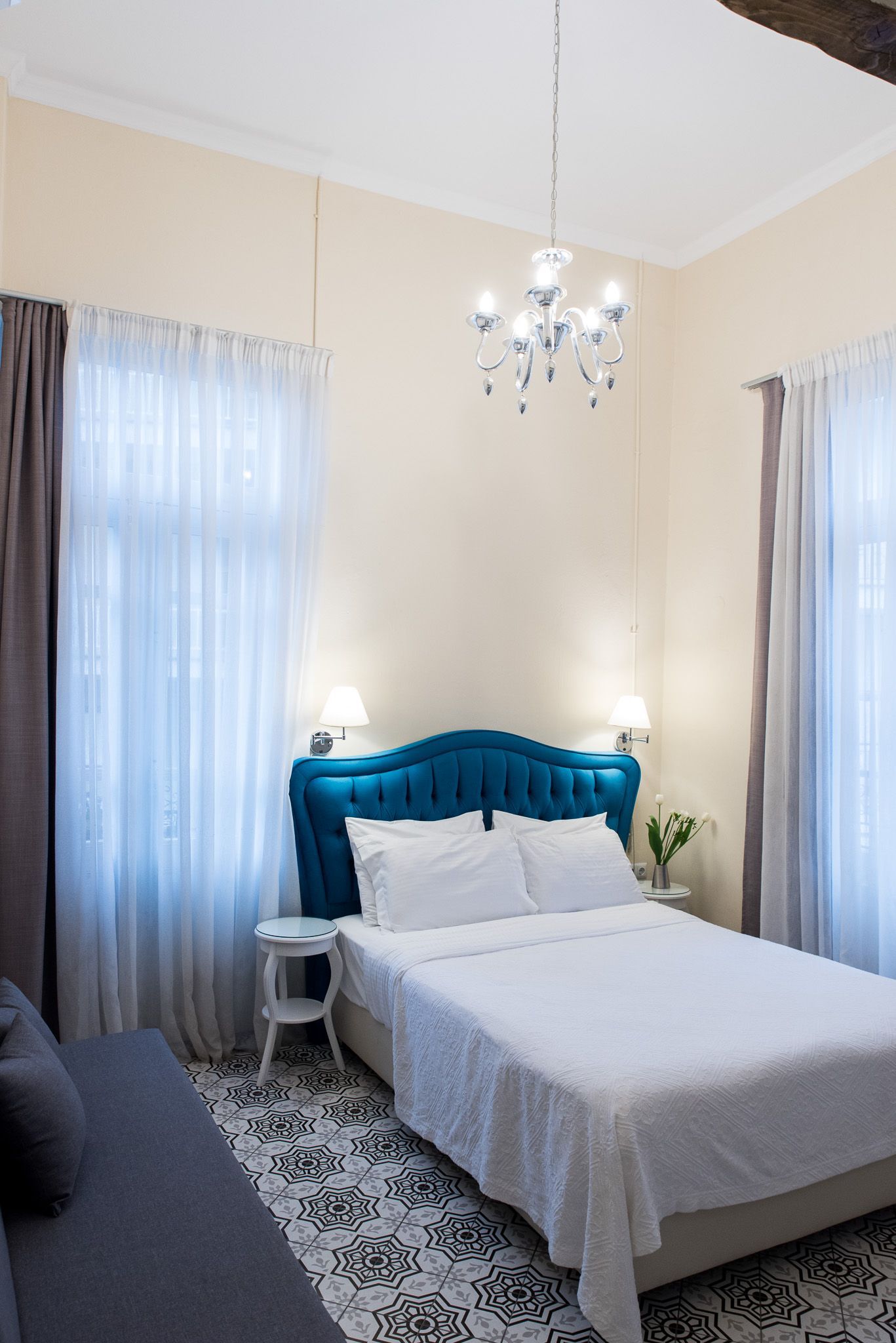A bedroom featuring a blue tufted headboard, white bedding, sheer curtains, and a crystal chandelier over patterned floors.