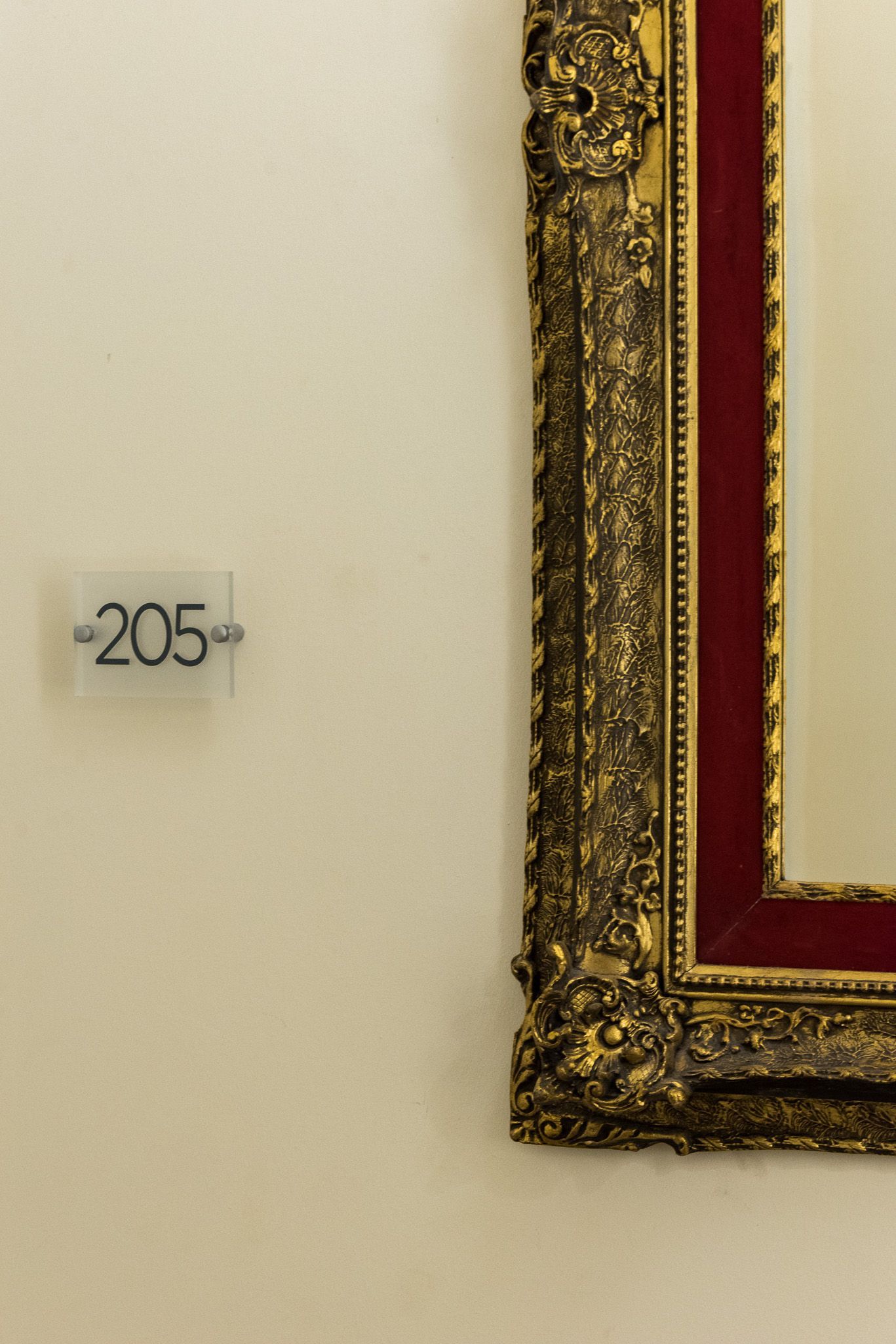 Μια μικρή λευκή τετράγωνη πινακίδα με την ένδειξη «205» σε έναν μπεζ τοίχο δίπλα σε μια μεγάλη, περίτεχνη χρυσή και κόκκινη κορνίζα.