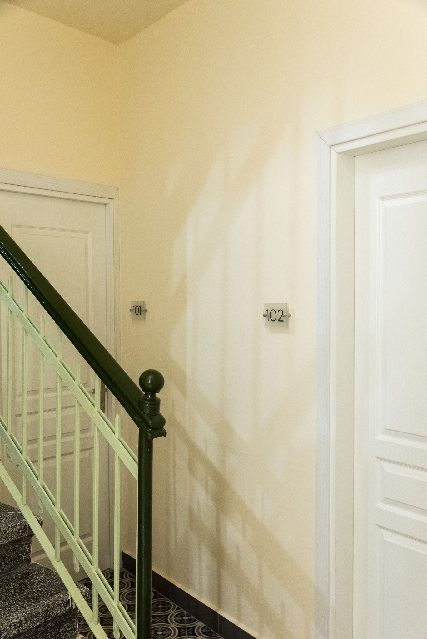 Μια σκάλα με σκούρο πράσινο κιγκλίδωμα οδηγεί δίπλα σε λευκές πόρτες σε έναν κίτρινο τοίχο με δύο διακόπτες φωτός.