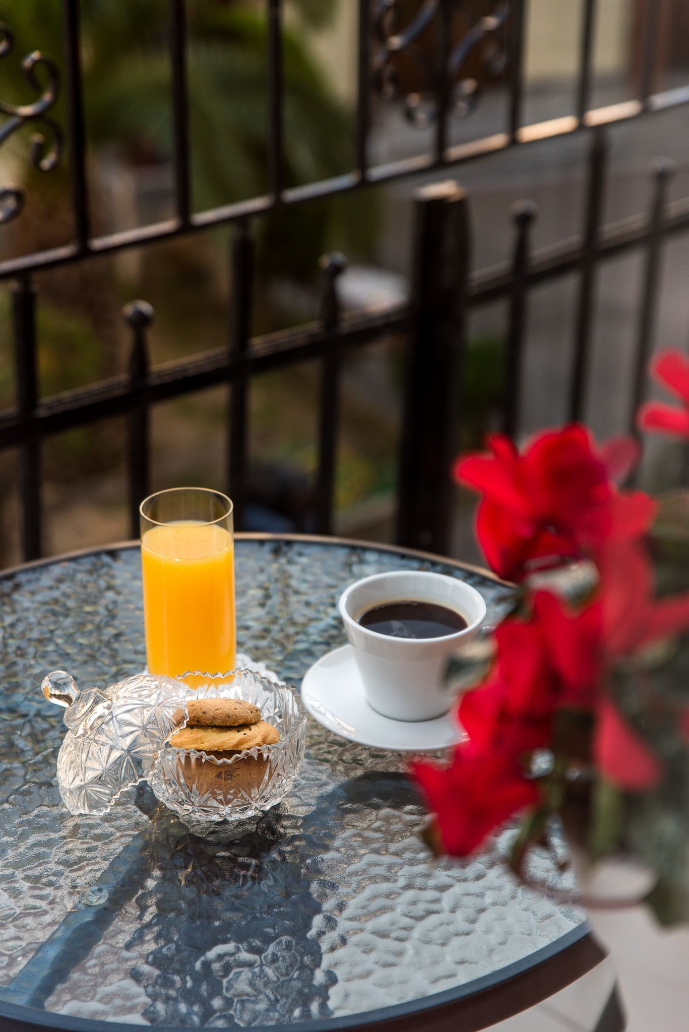 Ένα ποτήρι χυμό πορτοκάλι, καφές και μπισκότα σε ένα γυάλινο τραπέζι βεράντας, με κόκκινα λουλούδια στο προσκήνιο.