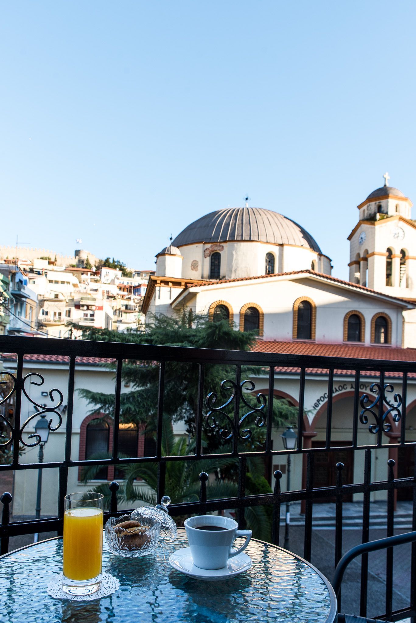 Ένα ποτήρι χυμό πορτοκάλι και ένα φλιτζάνι καφέ σε ένα τραπέζι μπαλκονιού, με μια εκκλησία και σπίτια στην πλαγιά του λόφου στο βάθος.