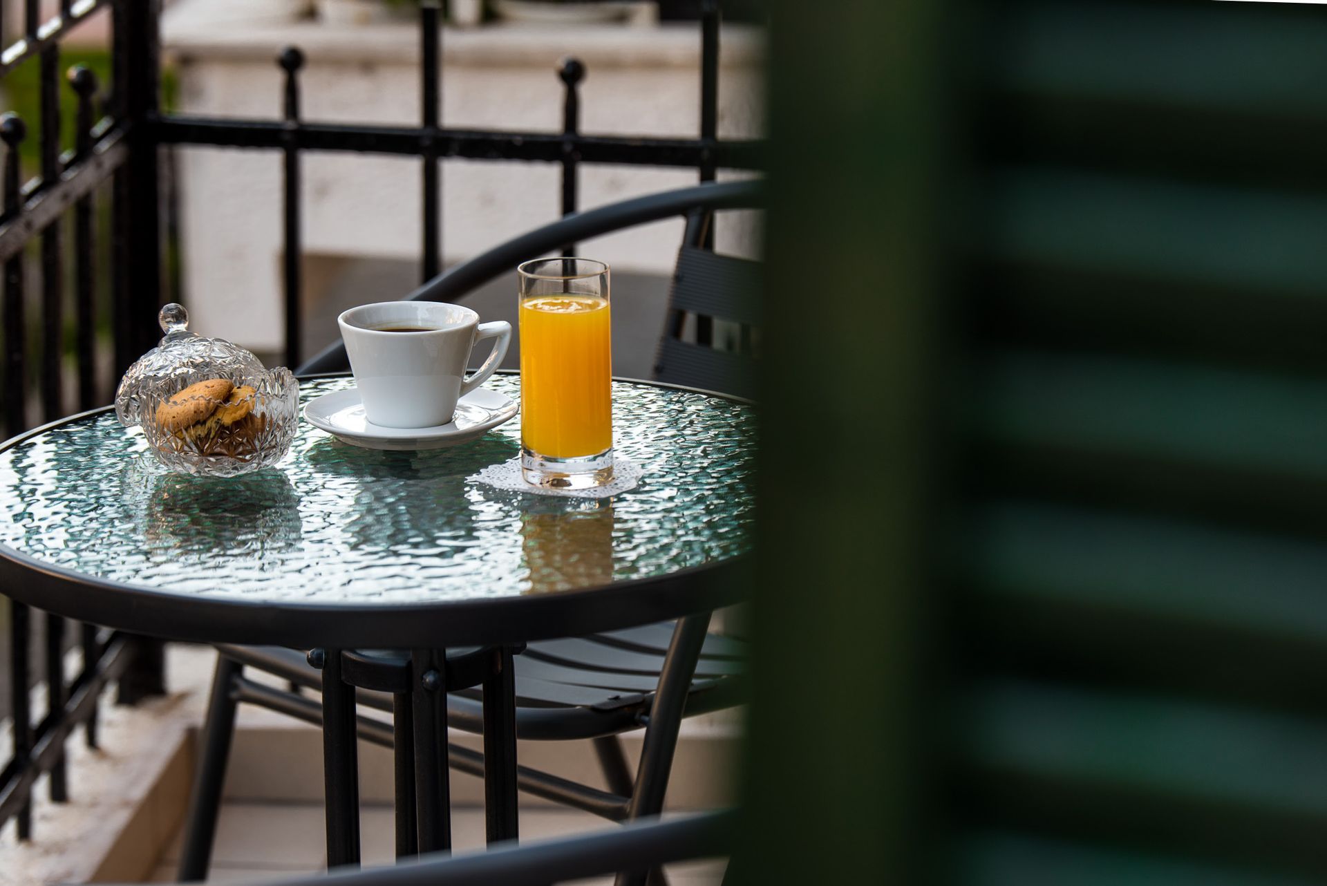 Ένα γυάλινο τραπέζι σε ένα μπαλκόνι περιέχει ένα ποτήρι χυμό πορτοκάλι, ένα φλιτζάνι καφέ και ένα μικρό διακοσμητικό μπολ με μπισκότα.