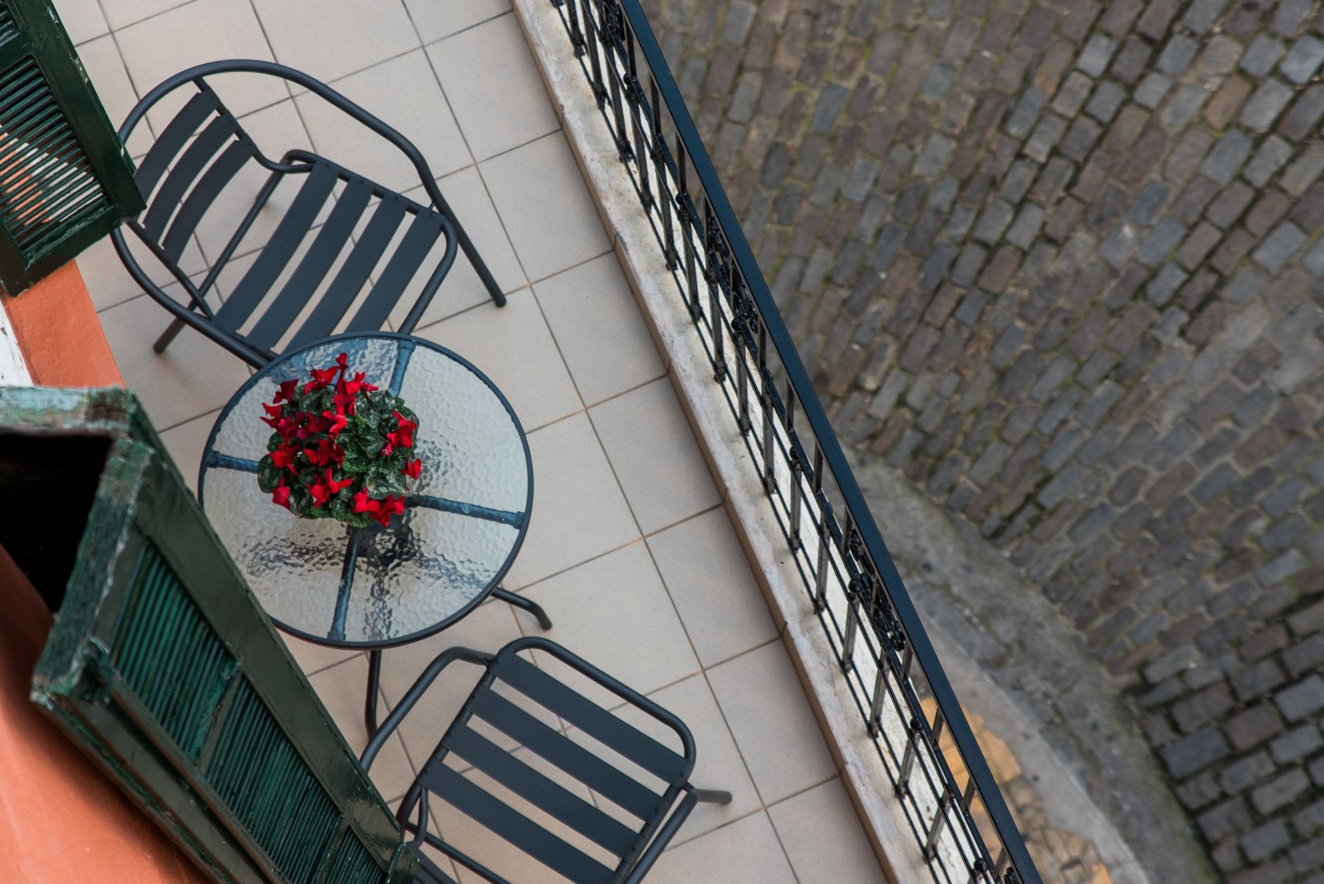 Δύο σκούρες μεταλλικές καρέκλες και ένα μικρό γυάλινο τραπέζι με ένα κόκκινο λουλουδάτο κεντρικό διακοσμητικό σε ένα μπαλκόνι με θέα σε έναν πλακόστρωτο δρόμο.