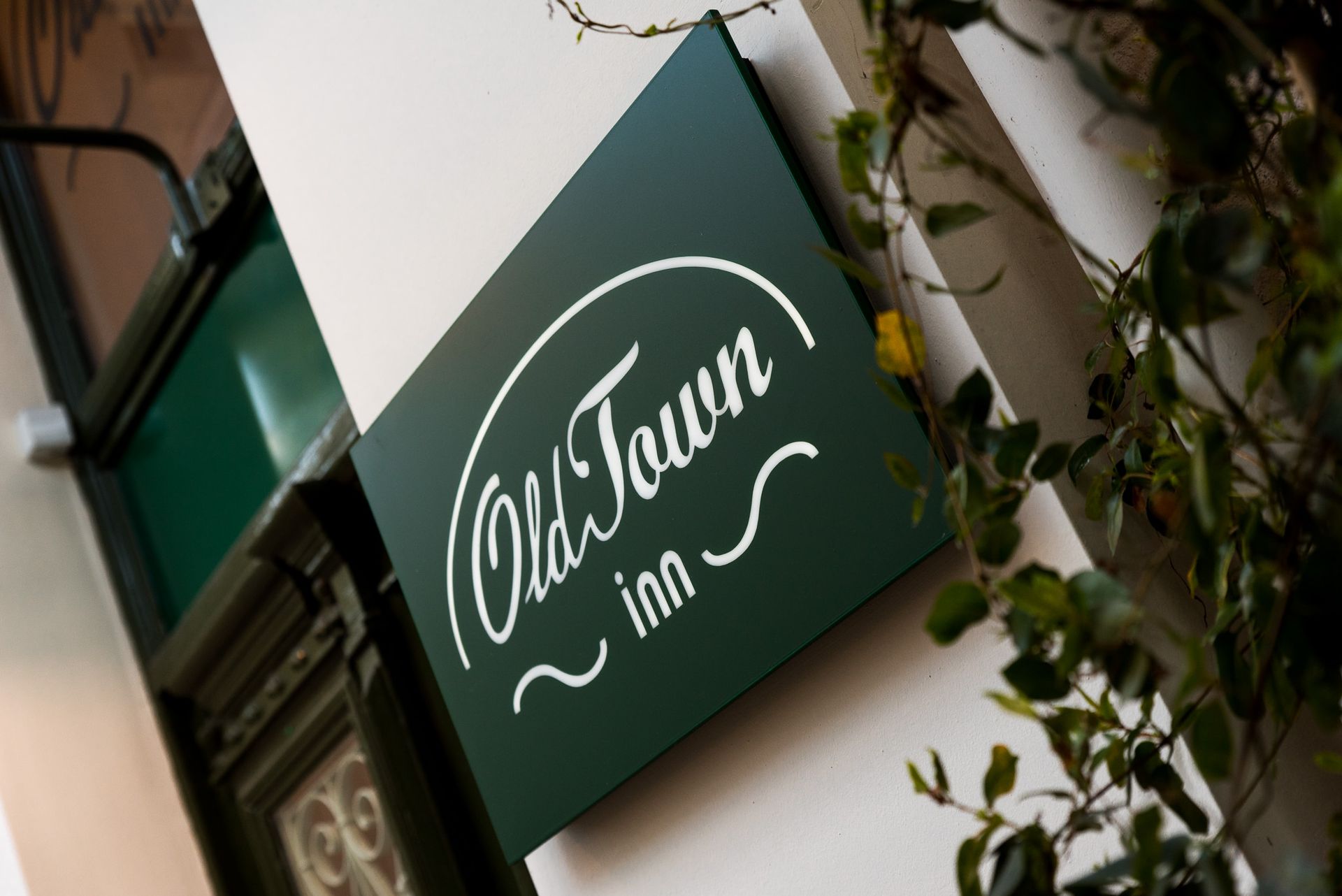 Μια πράσινη ορθογώνια πινακίδα με το λευκό στυλιζαρισμένο κείμενο «Old Town inn» τοποθετημένη σε έναν λευκό τοίχο δίπλα σε μια σκούρα πόρτα.