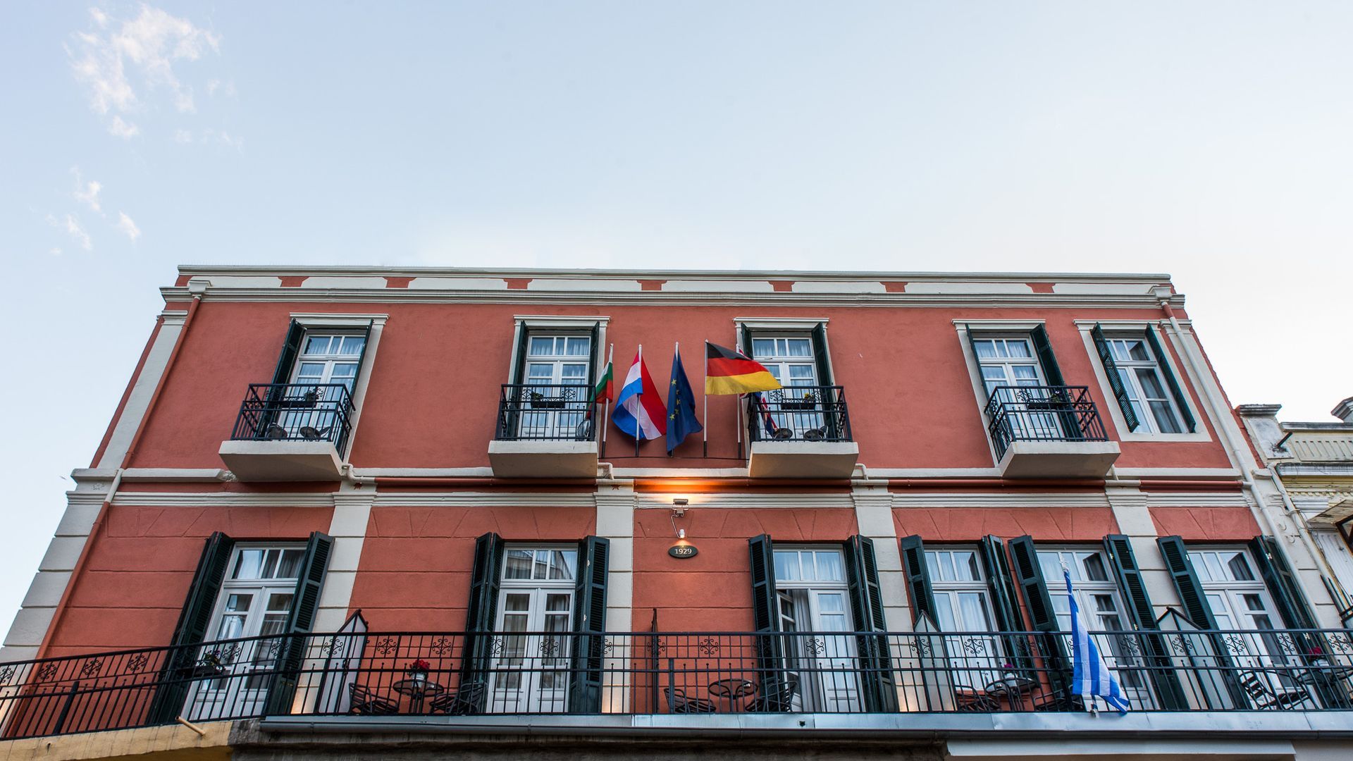 Ένα διώροφο κτίριο σε ροδακινί χρώμα με σκούρα παράθυρα, μπαλκόνια και ευρωπαϊκές σημαίες να κυματίζουν από πάνω.