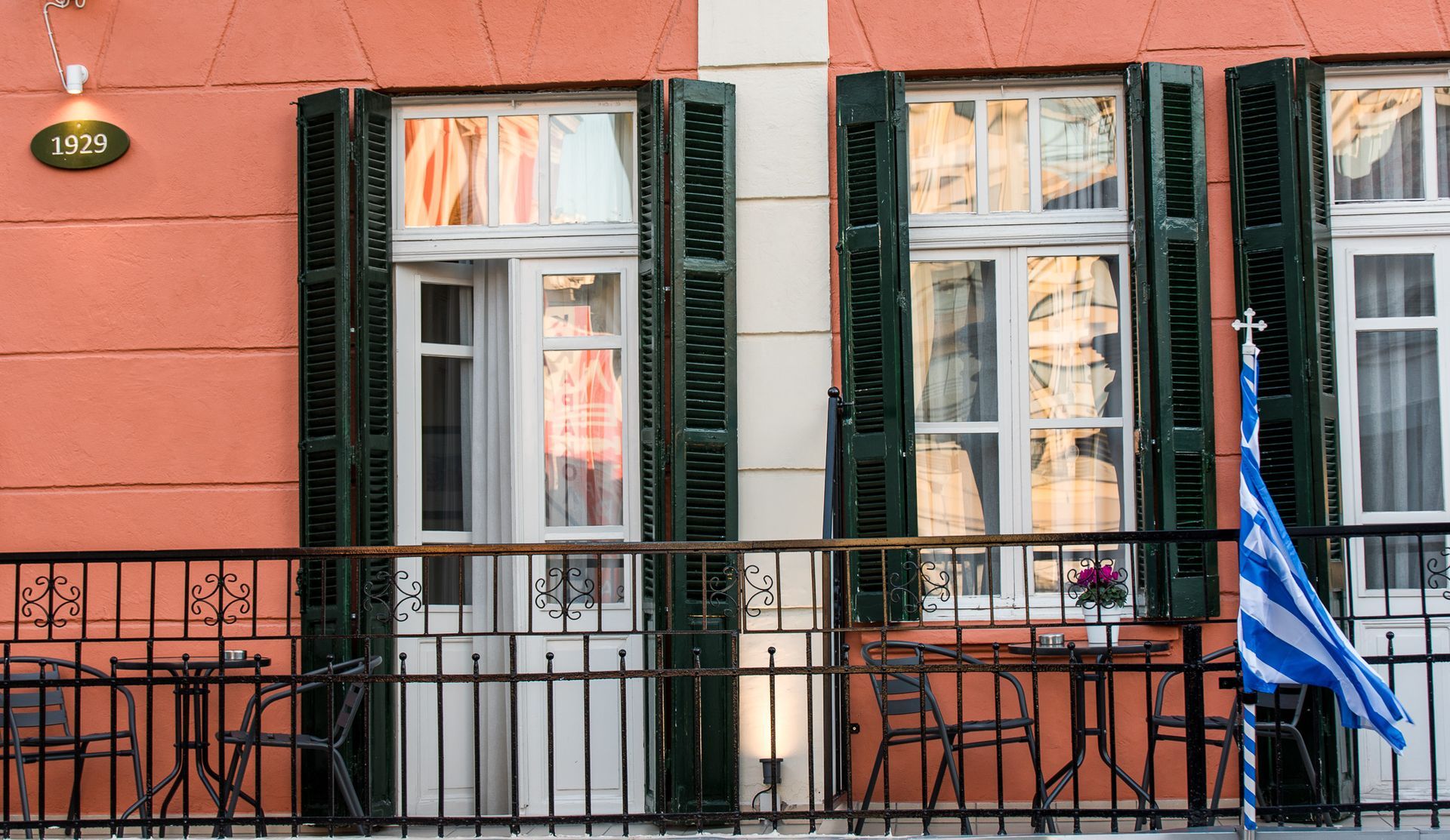 Ένα κτίριο σε σομόν χρώμα στο εξωτερικό του με δύο παράθυρα, σκούρα πράσινα παντζούρια, ένα μαύρο μεταλλικό κιγκλίδωμα και μια ελληνική σημαία.