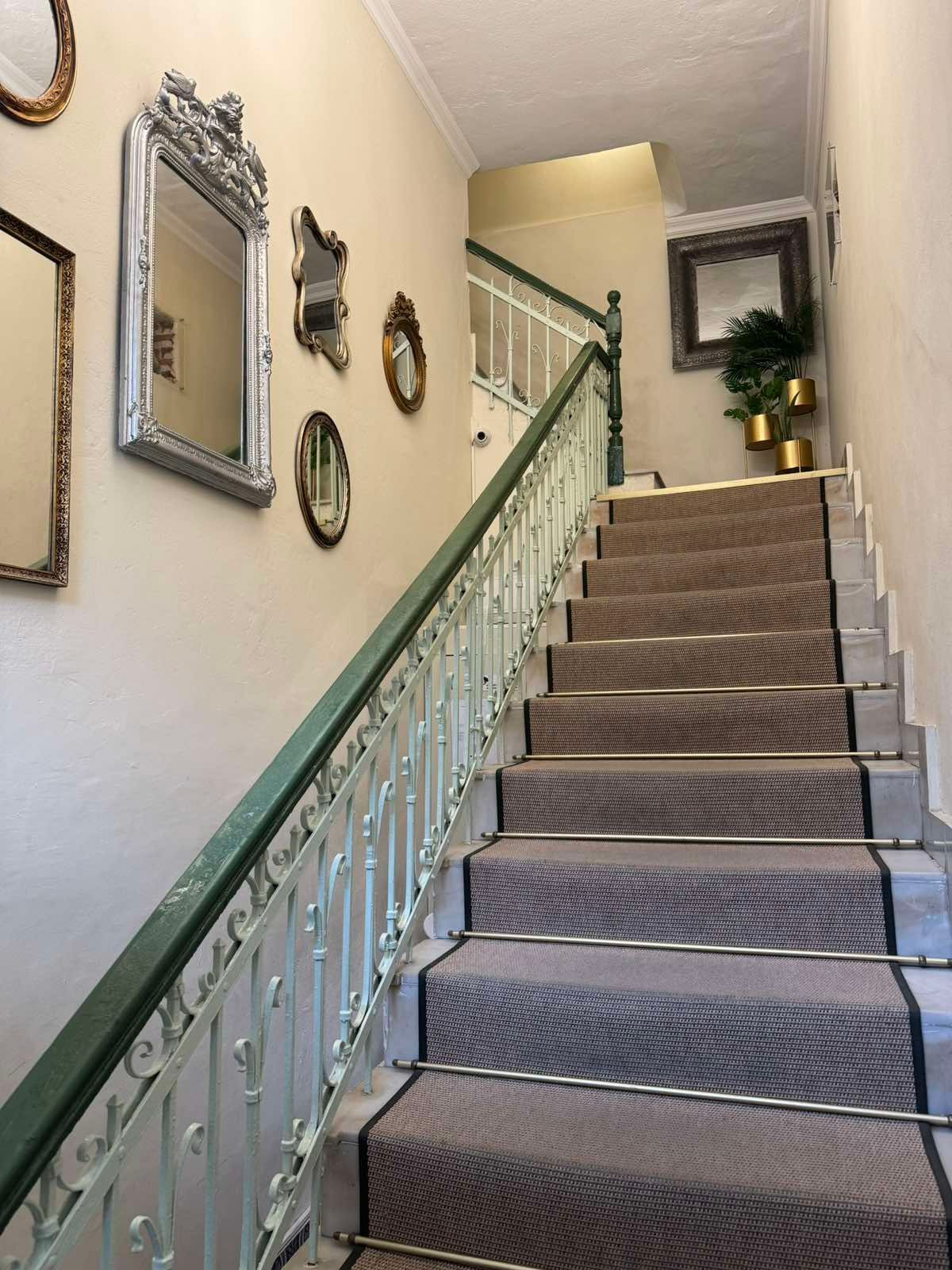 Σκαλοπάτια με γκρι δρομέα με σχέδια και πράσινο σιδερένιο κιγκλίδωμα, που ανηφορίζουν περνώντας από μια επιτοίχια στοά με διάφορους πλαισιωμένους καθρέφτες.