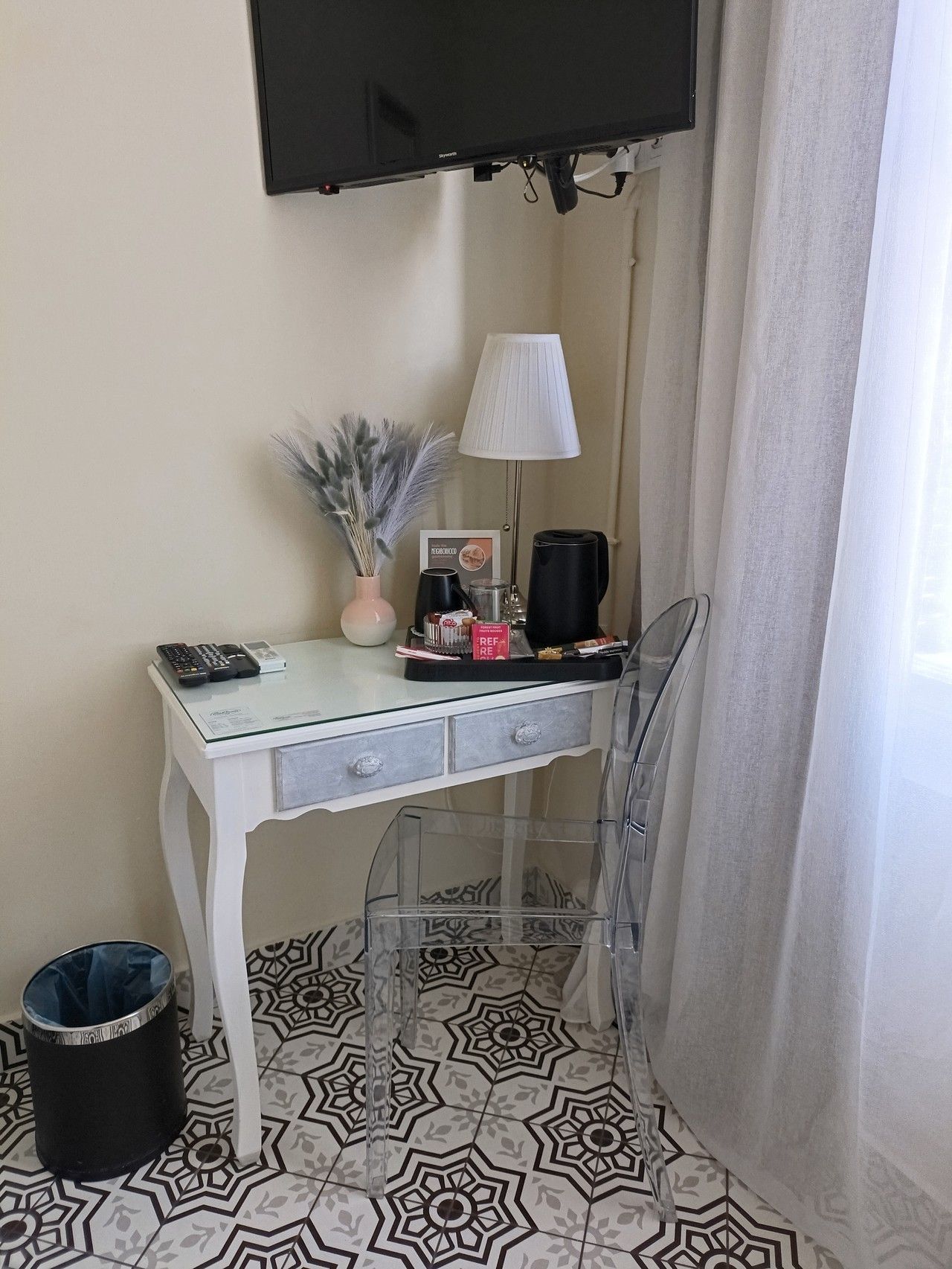 Ένα μικρό λευκό γραφείο νιπτήρα με συρτάρια, ένα φωτιστικό, ένα βάζο και ένα σετ τσαγιού, δίπλα σε μια διαφανή καρέκλα και ένα δάπεδο με σχέδια.