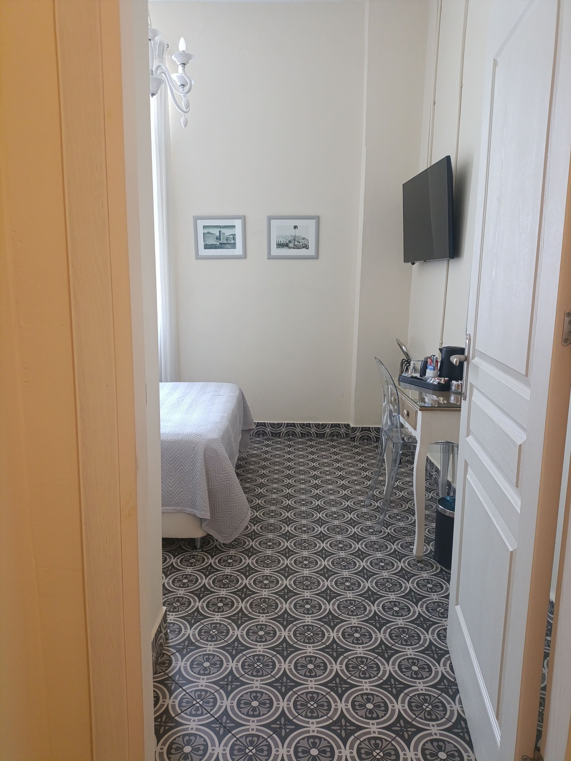 Θέα μέσα από μια πόρτα σε ένα μικρό δωμάτιο ξενοδοχείου με πλακάκια στο δάπεδο με σχέδια, ένα κρεβάτι, ένα γραφείο και μια επιτοίχια τηλεόραση.