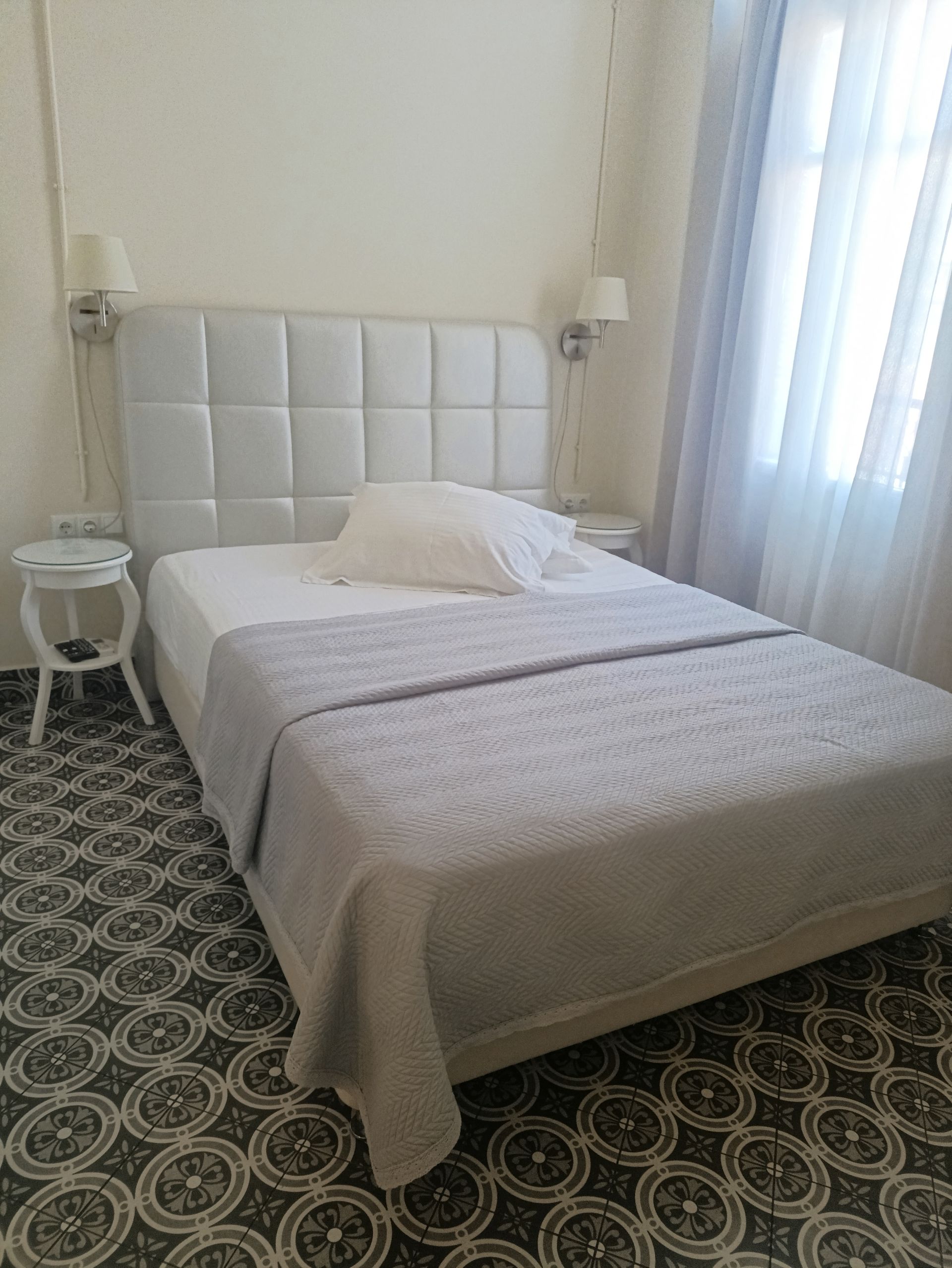 Ένα υπνοδωμάτιο με κεφαλάρι με φουντωτό γκρι χρώμα, λευκά κλινοσκεπάσματα, μια γκρι κουβέρτα και δύο μικρά λευκά κομοδίνα σε πλακάκια δαπέδου.