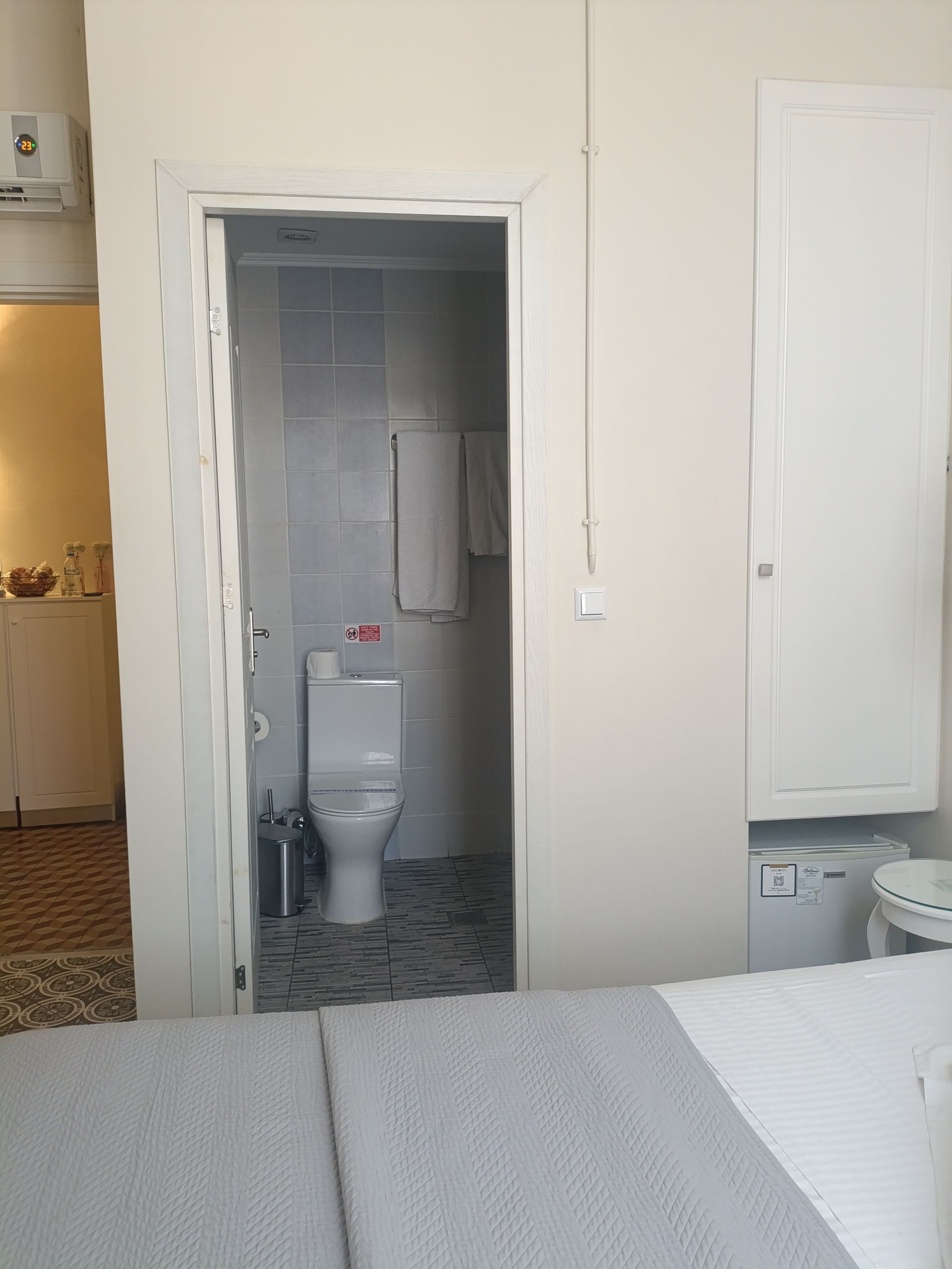 Μια άποψη υπνοδωματίου που δείχνει μια ανοιχτή πόρτα σε ένα μπάνιο με λευκή τουαλέτα και γκρι πλακάκια στο δάπεδο, δίπλα σε ένα λευκό ντουλάπι.