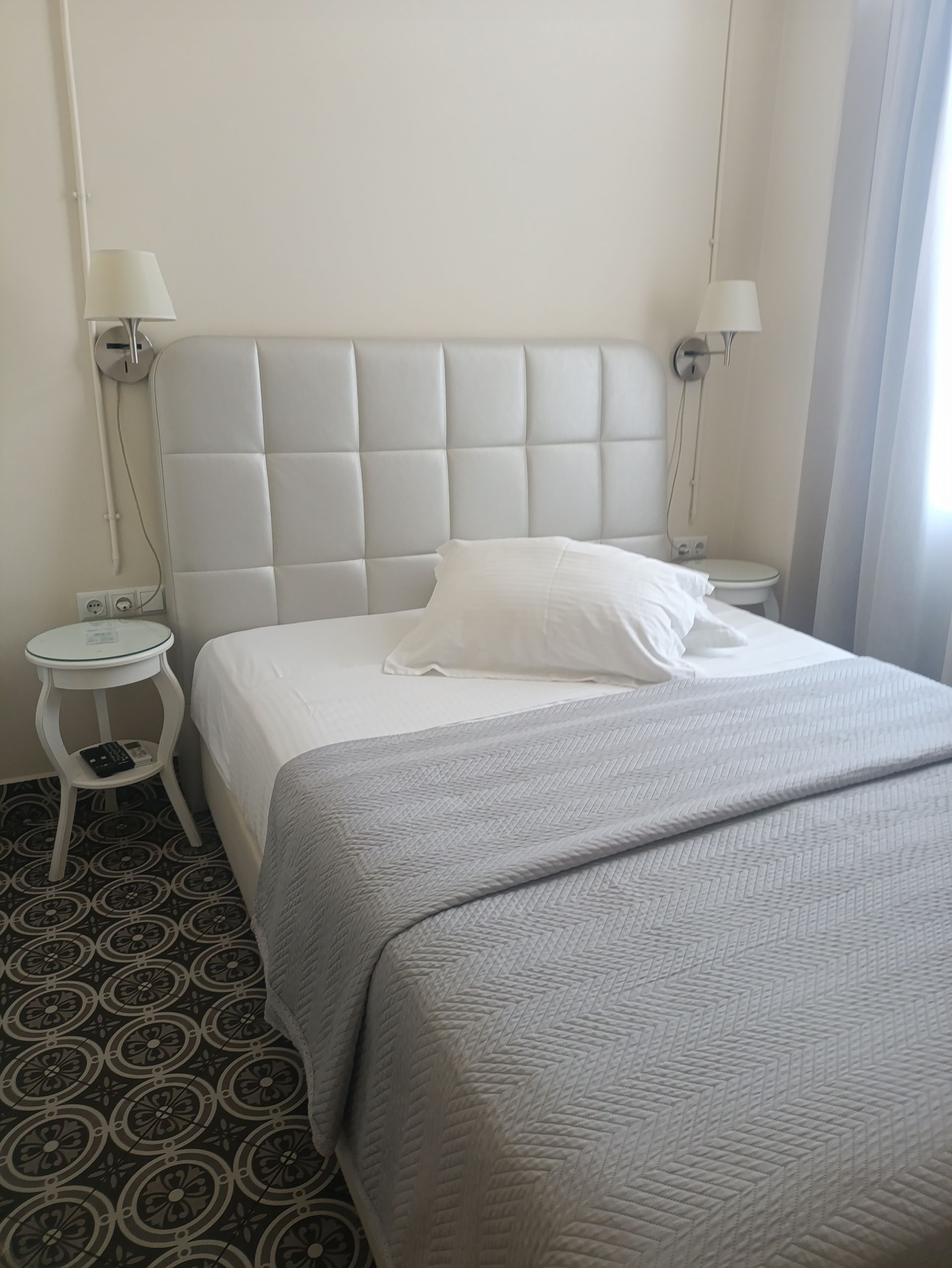 Ένα άνετο υπνοδωμάτιο ξενοδοχείου με λευκό καπιτονέ κεφαλάρι, μια γκρι πλεκτή κουβέρτα και ένα μικρό βοηθητικό τραπεζάκι πάνω σε ένα χαλί με σχέδια.