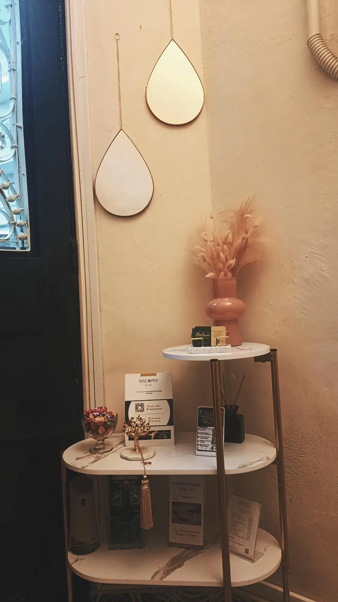 Δύο επιτοίχιοι καθρέφτες σε σχήμα δακρύου κρέμονται πάνω από ένα τριώροφο βοηθητικό τραπεζάκι από λευκό μάρμαρο και χρυσό μέταλλο με διάφορα διακοσμητικά.