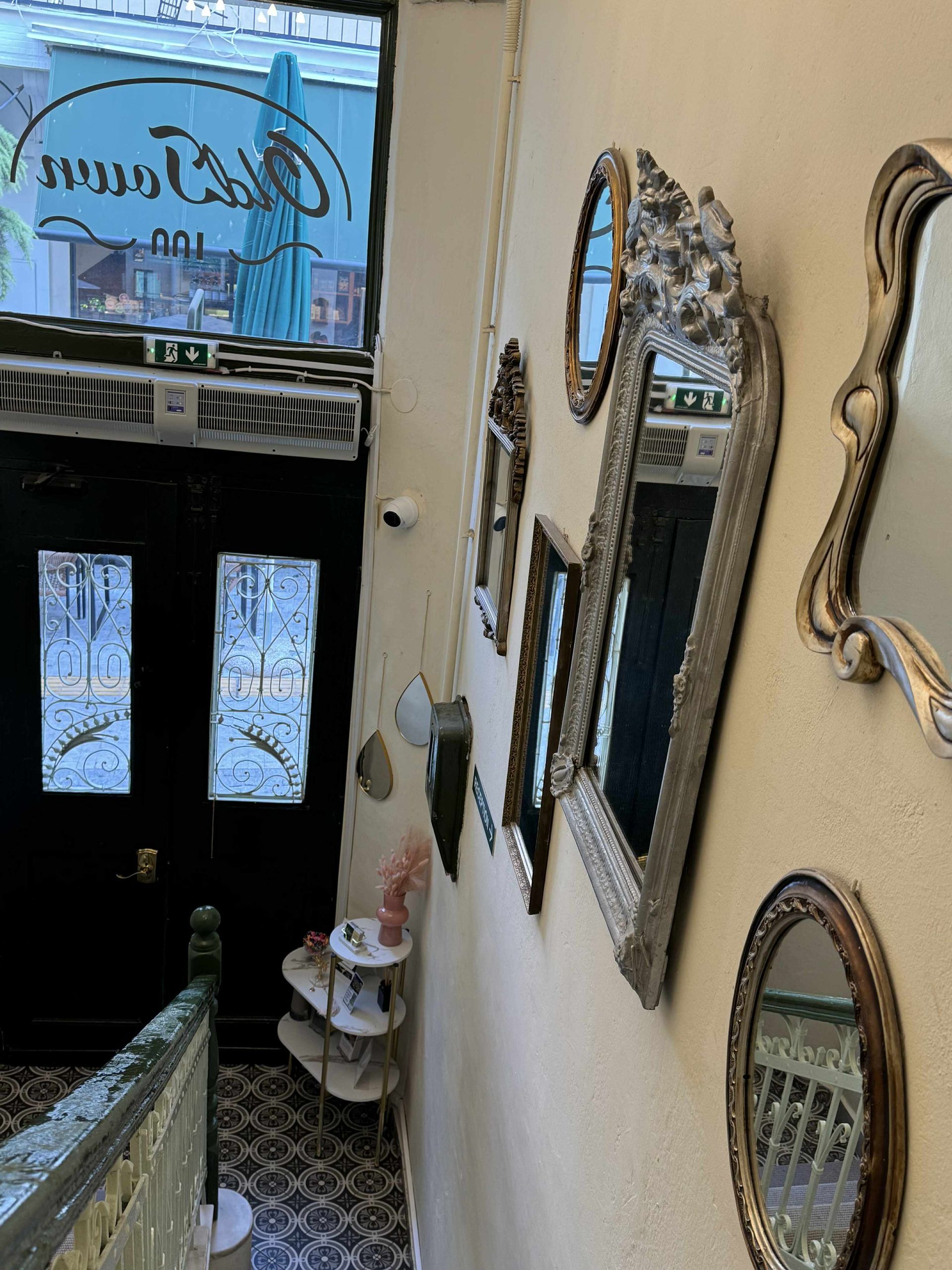 Ένα κλιμακοστάσιο με αρκετούς περίτεχνους καθρέφτες vintage κρεμασμένους σε έναν κρεμ τοίχο δίπλα σε μια πόρτα με σκούρο τζάμι.