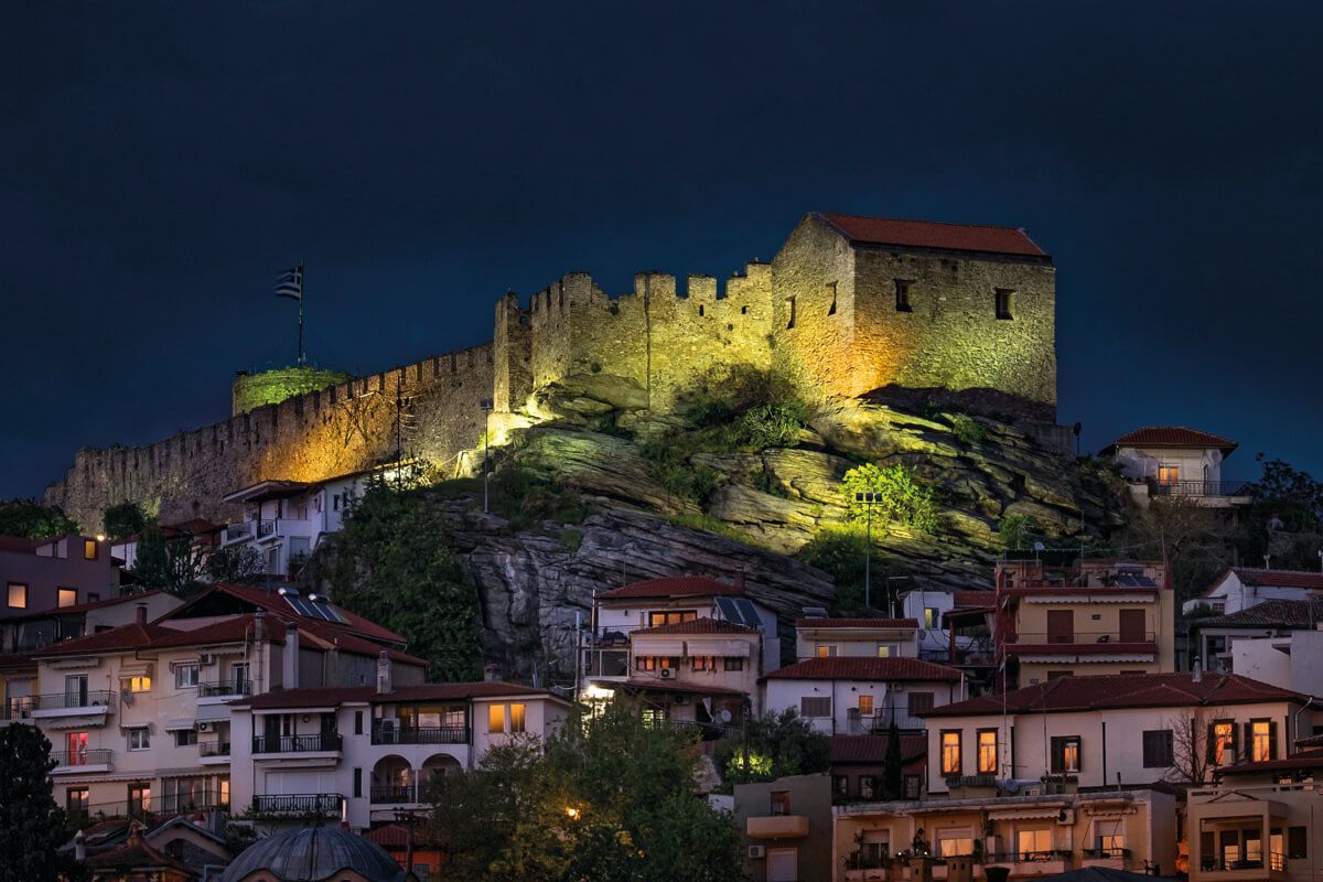 An illuminated medieval stone castle sits atop a rocky hill, overlooking a town of small buildings at twilight.