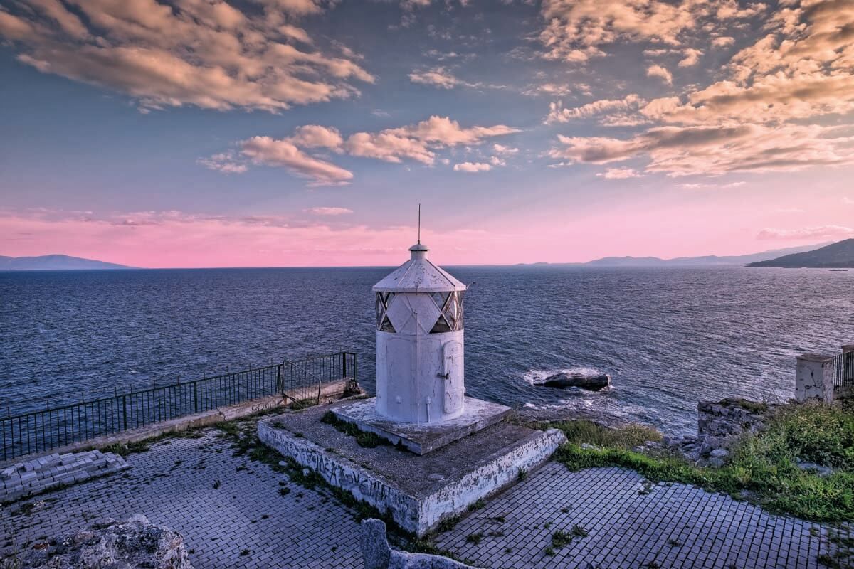 A white lighthouse stands on a stone cliff overlooking a calm sea under a sunset sky with pink and orange clouds.