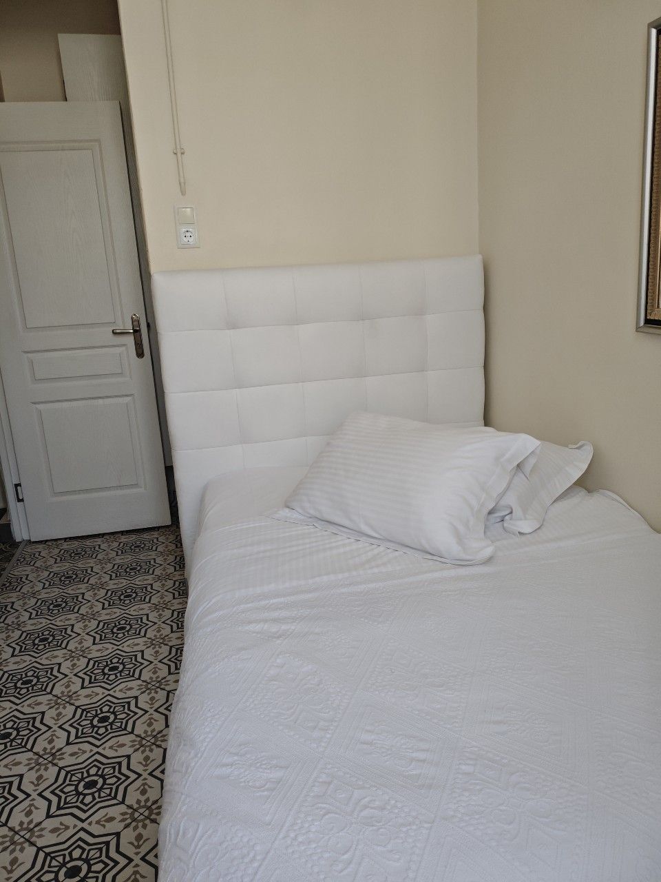 Ένα λευκό κρεβάτι με καπιτονέ κεφαλάρι και λευκά κλινοσκεπάσματα, τοποθετημένα δίπλα σε μια λευκή πόρτα σε ένα δωμάτιο με πλακάκια στο δάπεδο με σχέδια.