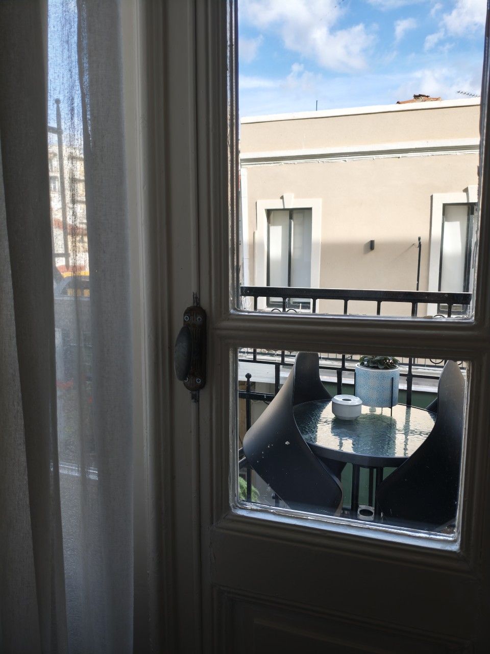 Μια θέα από το μπαλκόνι μέσα από ένα παράθυρο πόρτας δείχνει ένα μικρό στρογγυλό τραπέζι με δύο καρέκλες κάτω από έναν φωτεινό, μερικώς συννεφιασμένο ουρανό.
