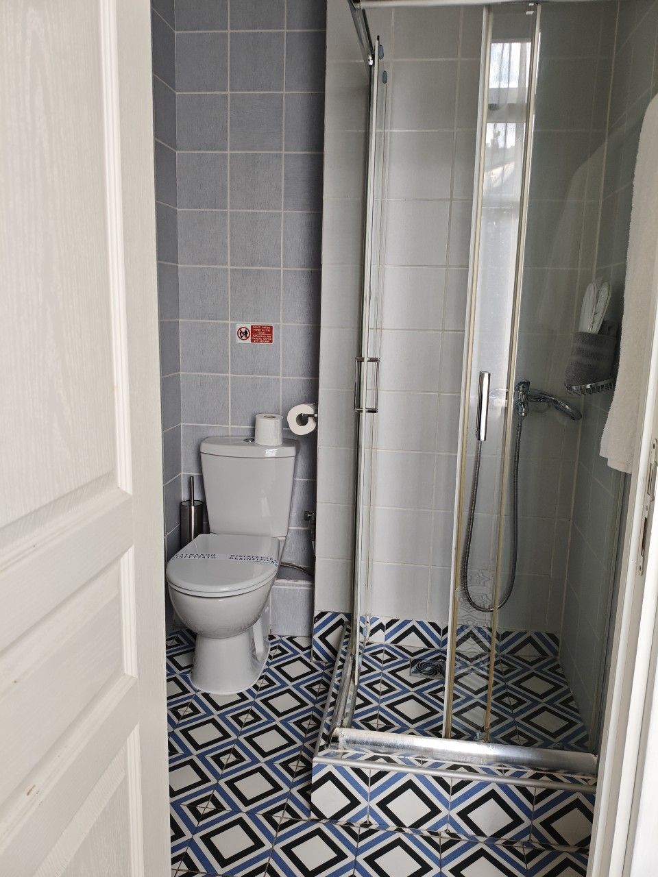 Ένα μικρό μπάνιο με λευκή τουαλέτα και γυάλινη καμπίνα ντους σε δάπεδο με μπλε και άσπρα πλακάκια με σχέδια.