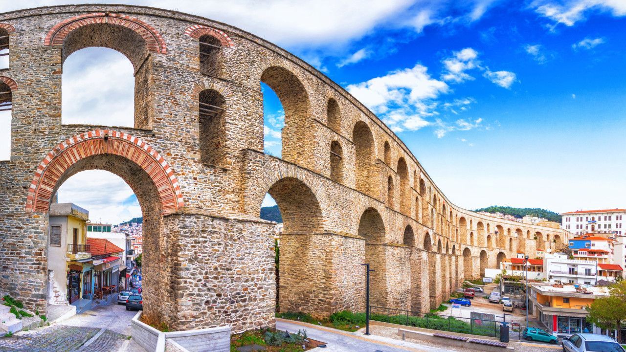 Kavala Aqueduct: A towering, ancient stone arched structure integrated into a modern Greek city under a sunny blue sky.