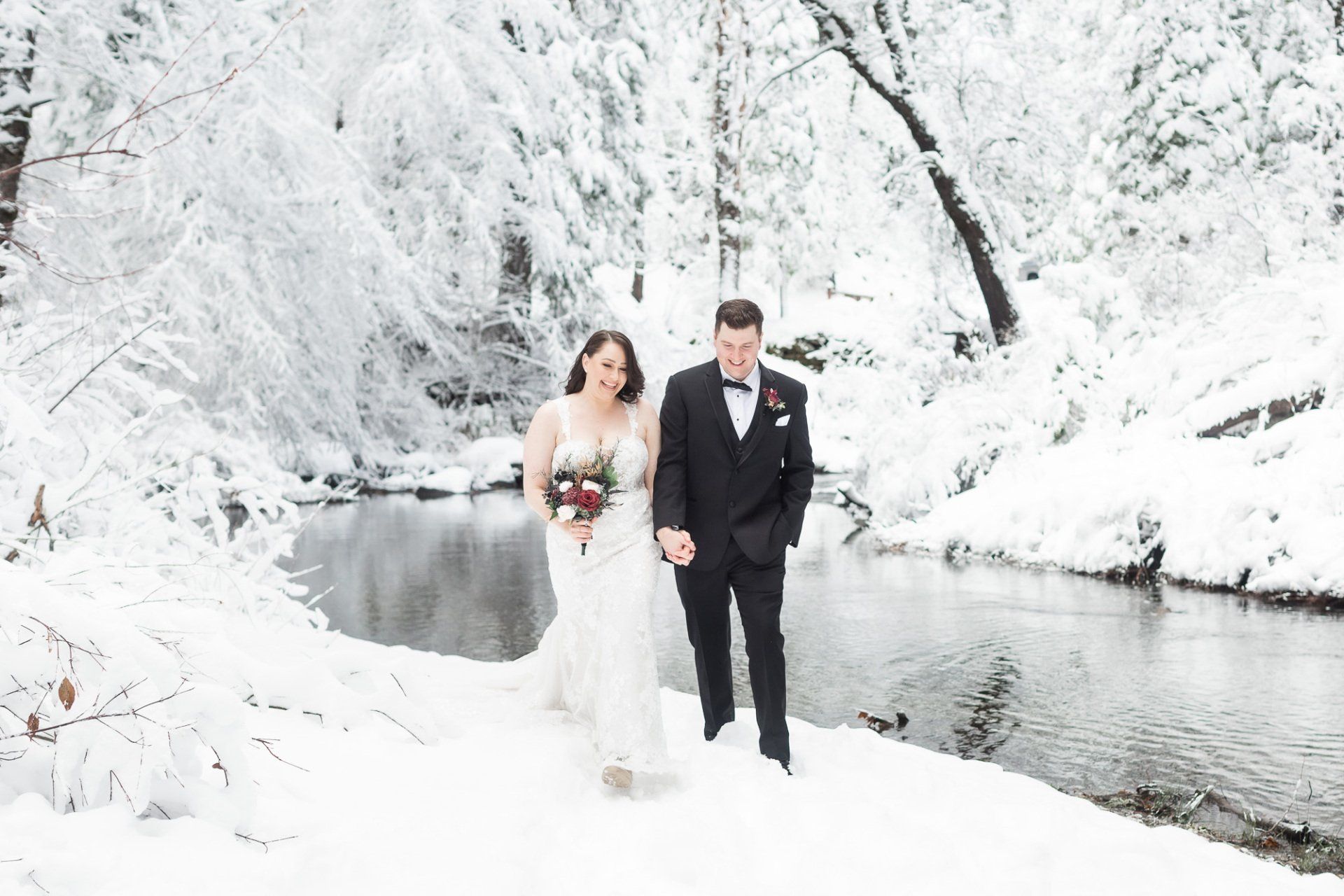 A bride and groom are standing next to a river in the snow.