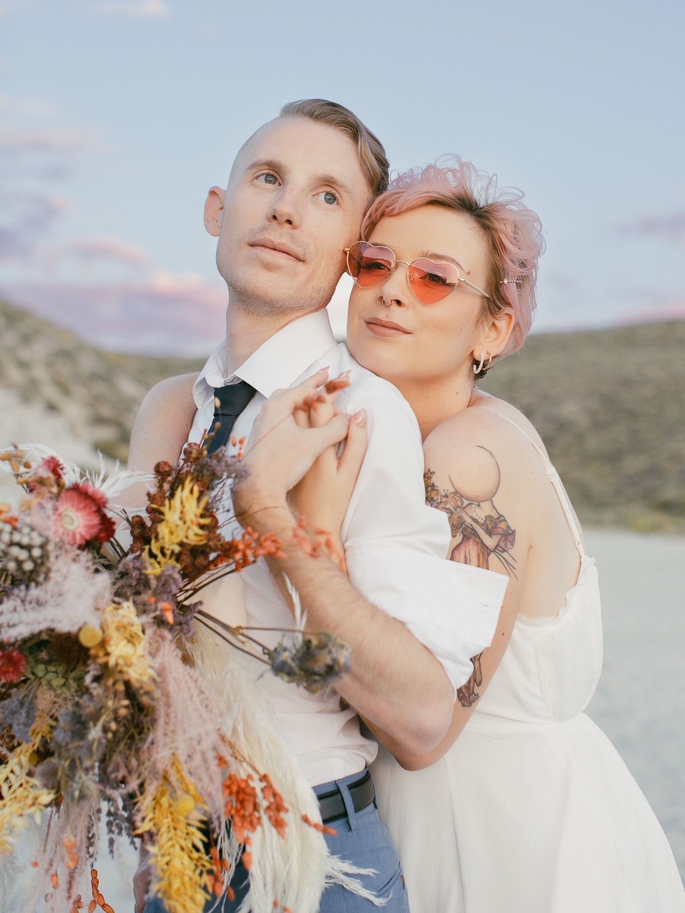 A bride and groom are posing for a picture on the beach . the bride is wearing pink sunglasses.