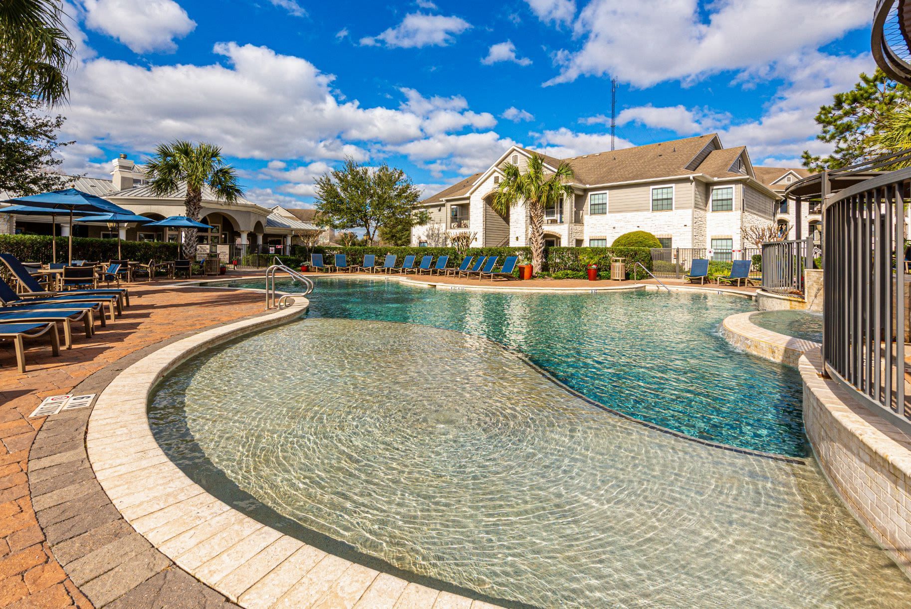 A large swimming pool surrounded by chairs and umbrellas in front of a building at Marquis Grand Lakes in Richmond, TX.