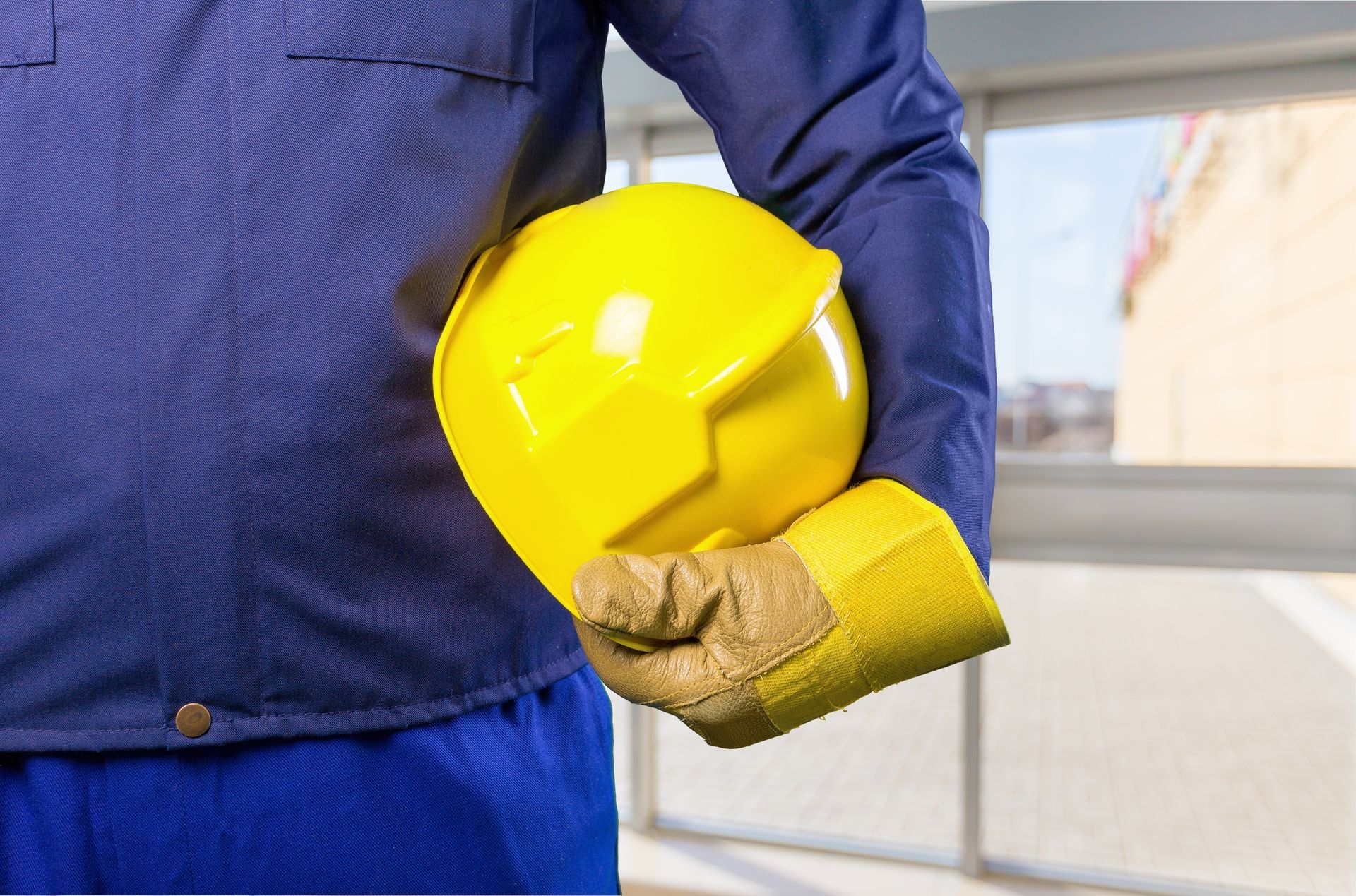 Worker in blue jumpsuit holding a yellow hard hat and wearing a yellow work glove.