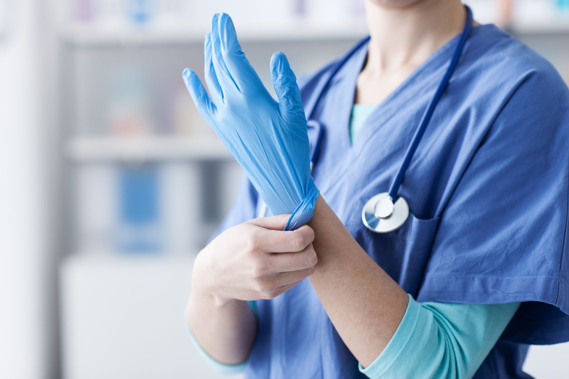 Medical professional in blue scrubs putting on a blue glove, stethoscope around neck.
