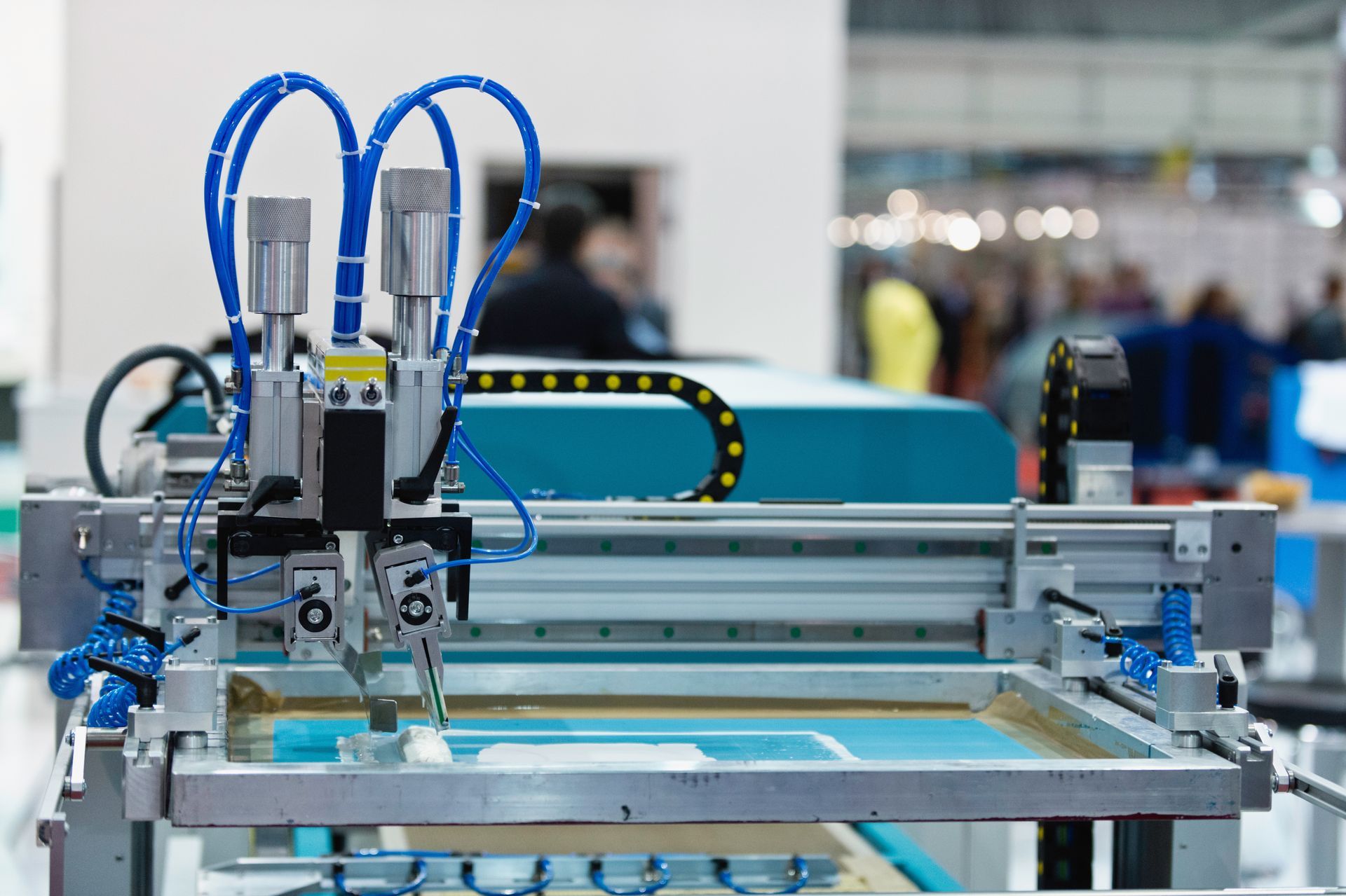 Screen printing machine with blue and silver components at an event, people blurred in the background.