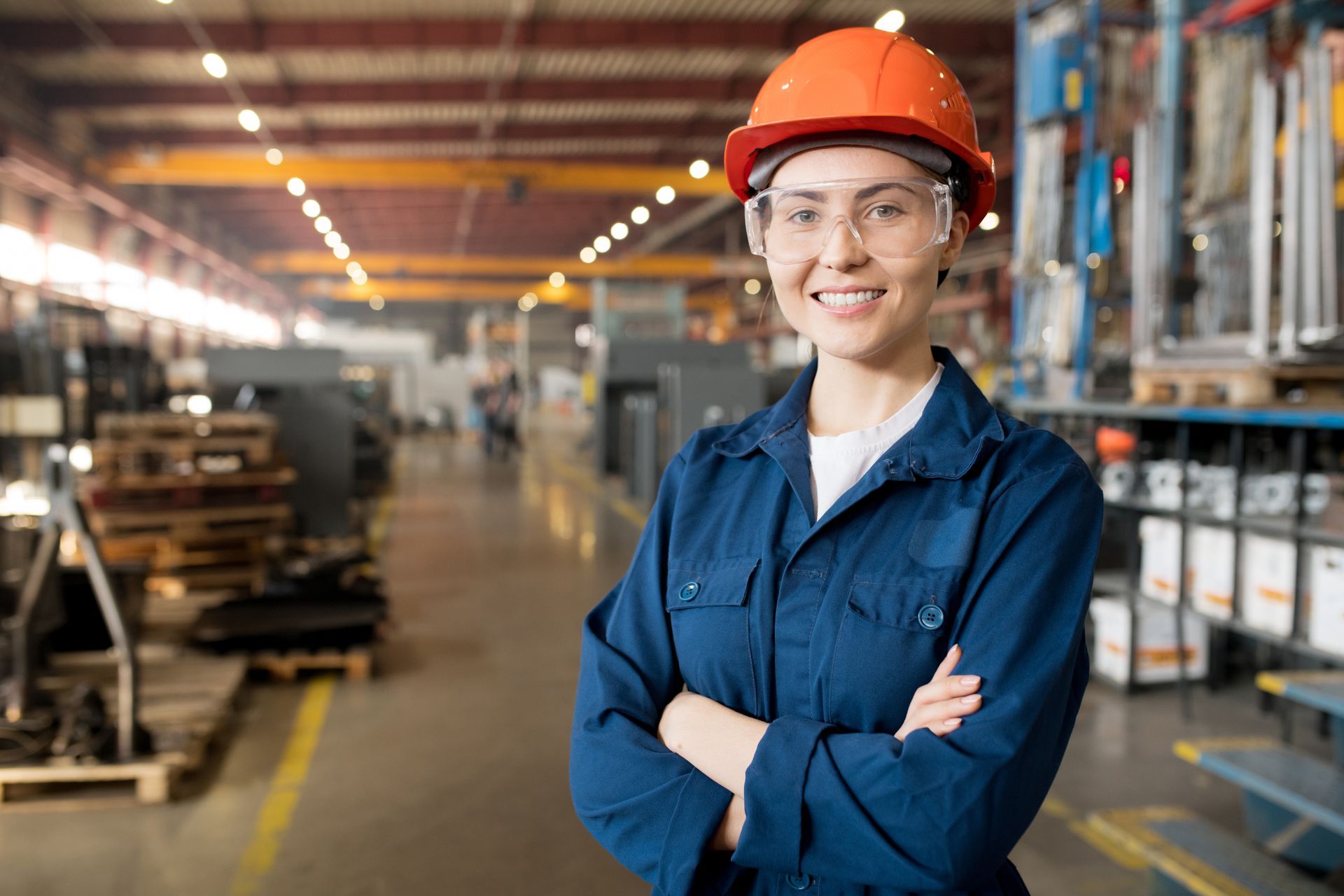 Woman in orange hard hat and safety glasses, arms crossed, smiling in a factory.
