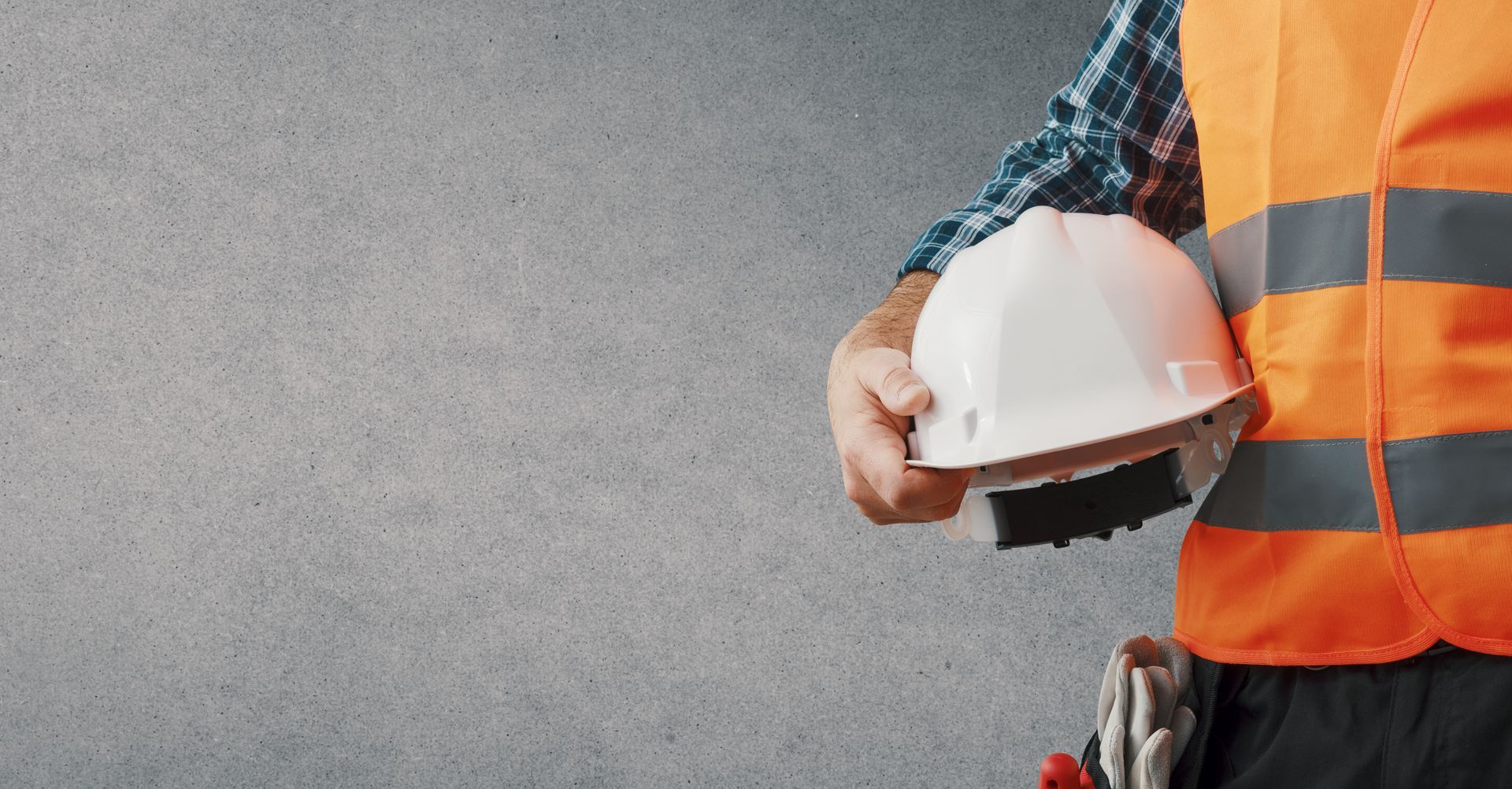 Construction worker holds white hard hat against a gray textured wall, wearing an orange safety vest.