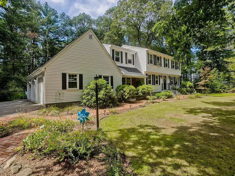 Two-story white house with black shutters, green lawn, trees, and blue pinwheel in front.