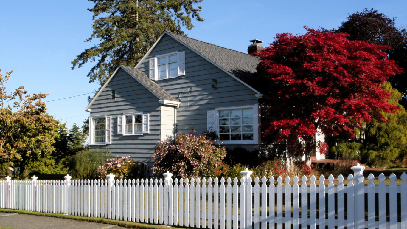 Gray house behind a white picket fence, vibrant red tree in the fall season.